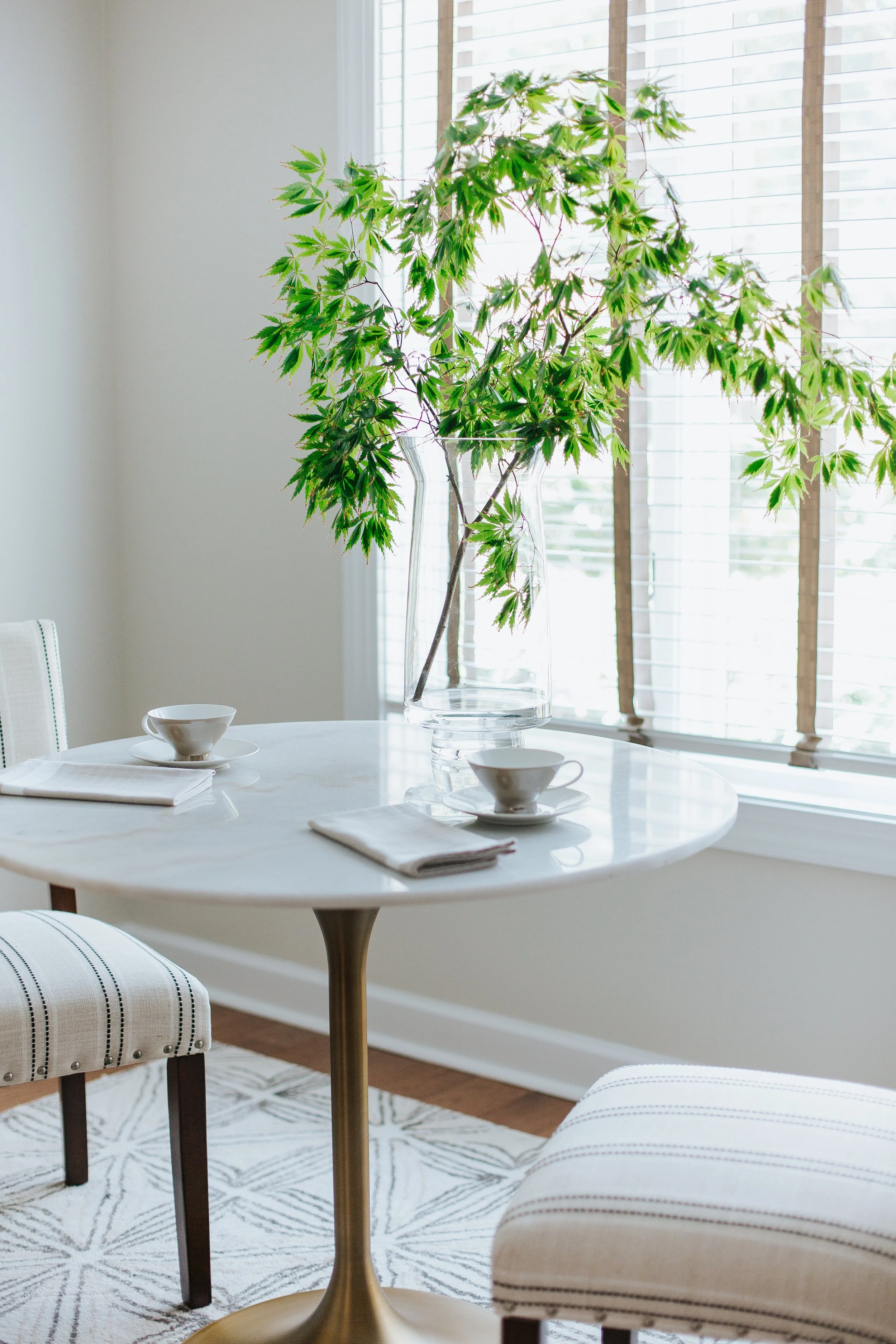 A round white dining table with two white teacups and a cloth napkin, and a tall glass vase with green maple leaves, near large windows with blinds, in a bright room with a patterned rug.