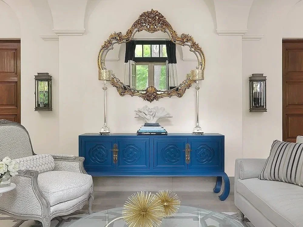 Decorative living room with a large ornate mirror, blue cabinet, white patterned armchair, striped sofa and wall sconces.