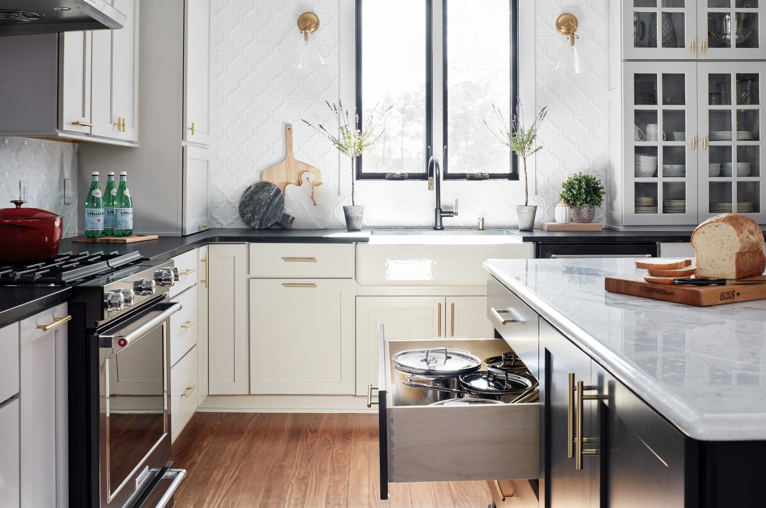 A bright kitchen with white cabinets, a black countertop, and a large window above the sink. There are plants and decorative items on the counter, a loaf of bread on a cutting board, and a stove on the left side.