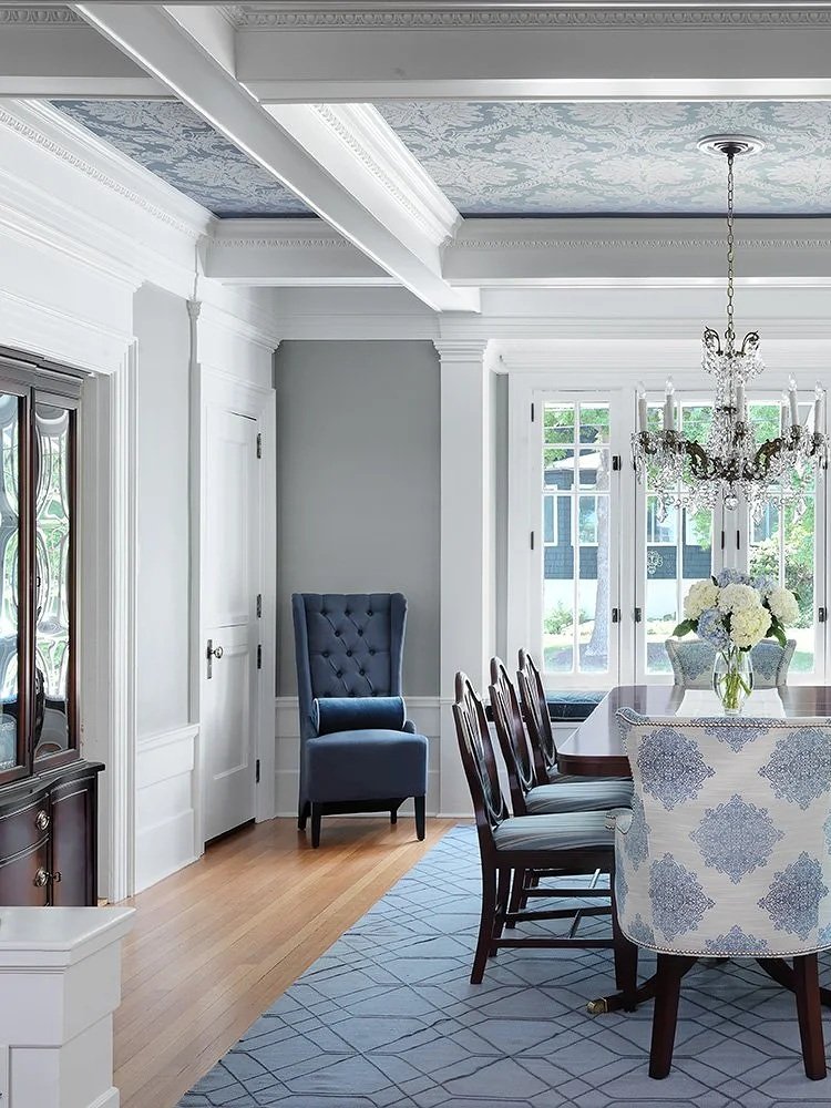 A dining room with a chandelier, large window with white framing, a wooden dining table with highback patterned chairs, a china cabinet, and a patterned rug. The ceiling features decorative molding and painted designs.