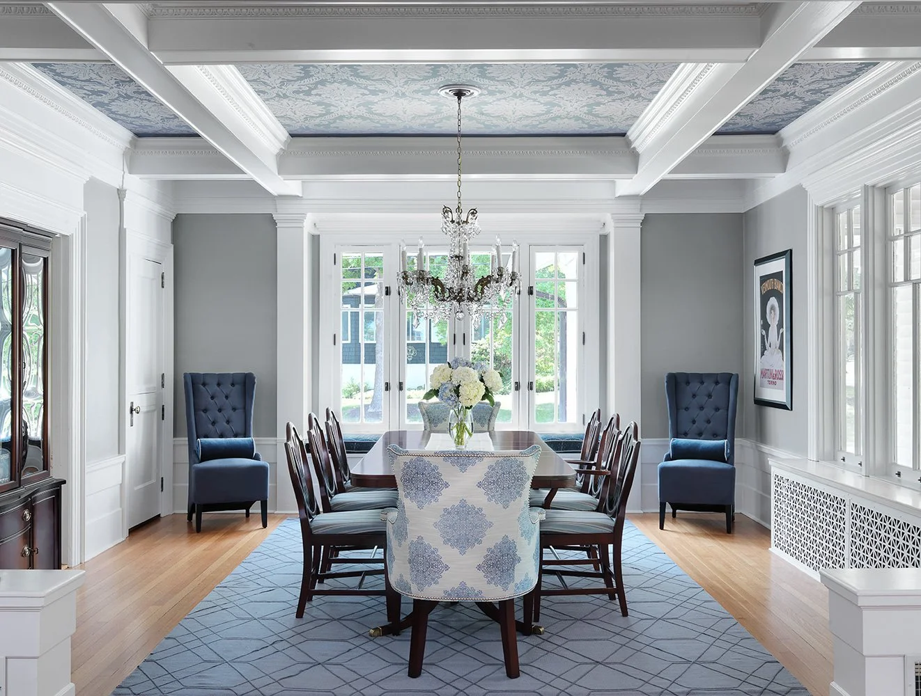 Elegant dining room with a chandelier, a large wooden dining table, upholstered armchairs, and large windows with white trim, in a classic style.