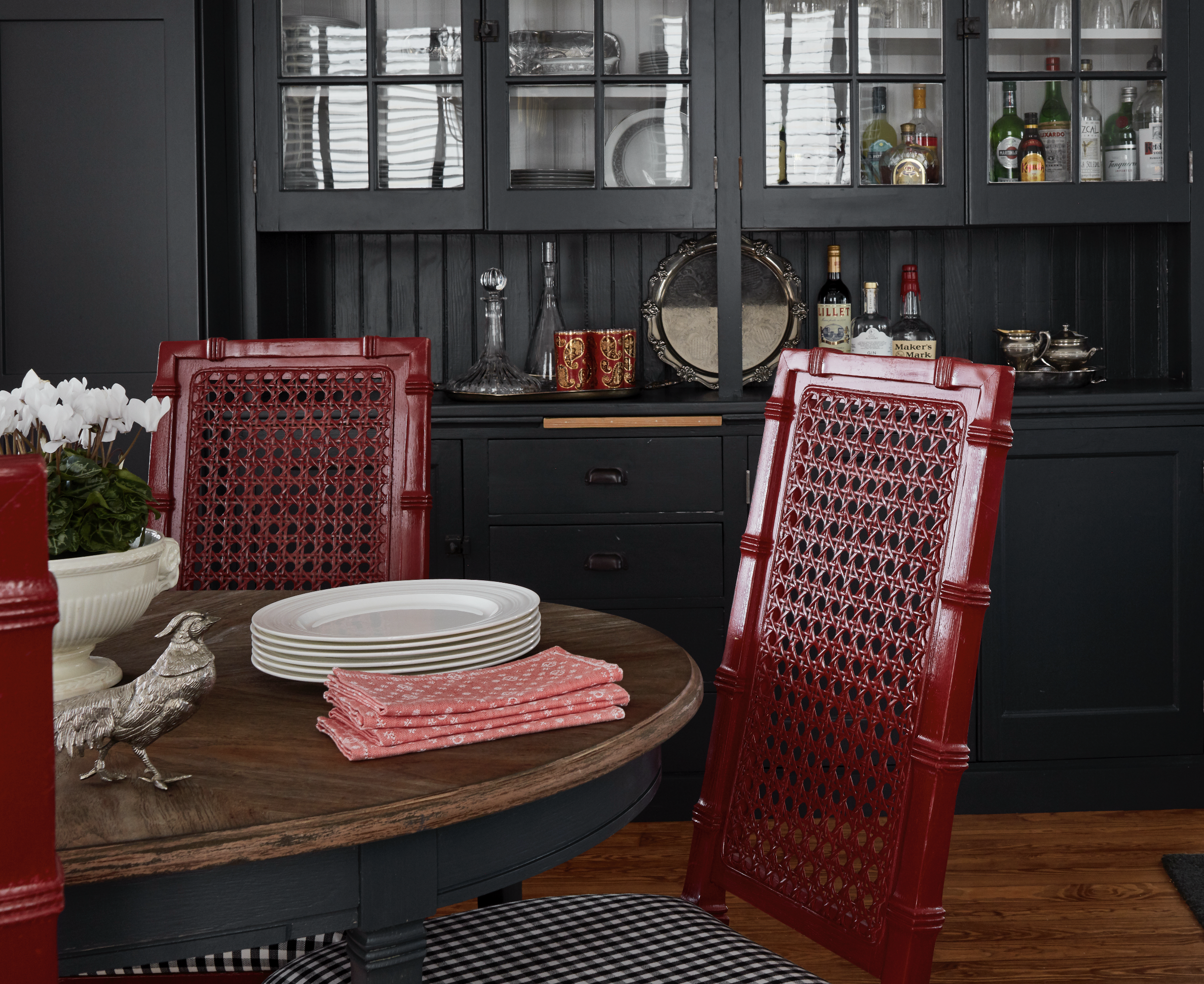 A table with a stack of white plates, a silver ornamental elephant, and red napkins. Red chairs with intricate back designs surround the table. Behind, a black wood cabinet with glass doors displays various bottles and decorative items, with reflections of window blinds visible.