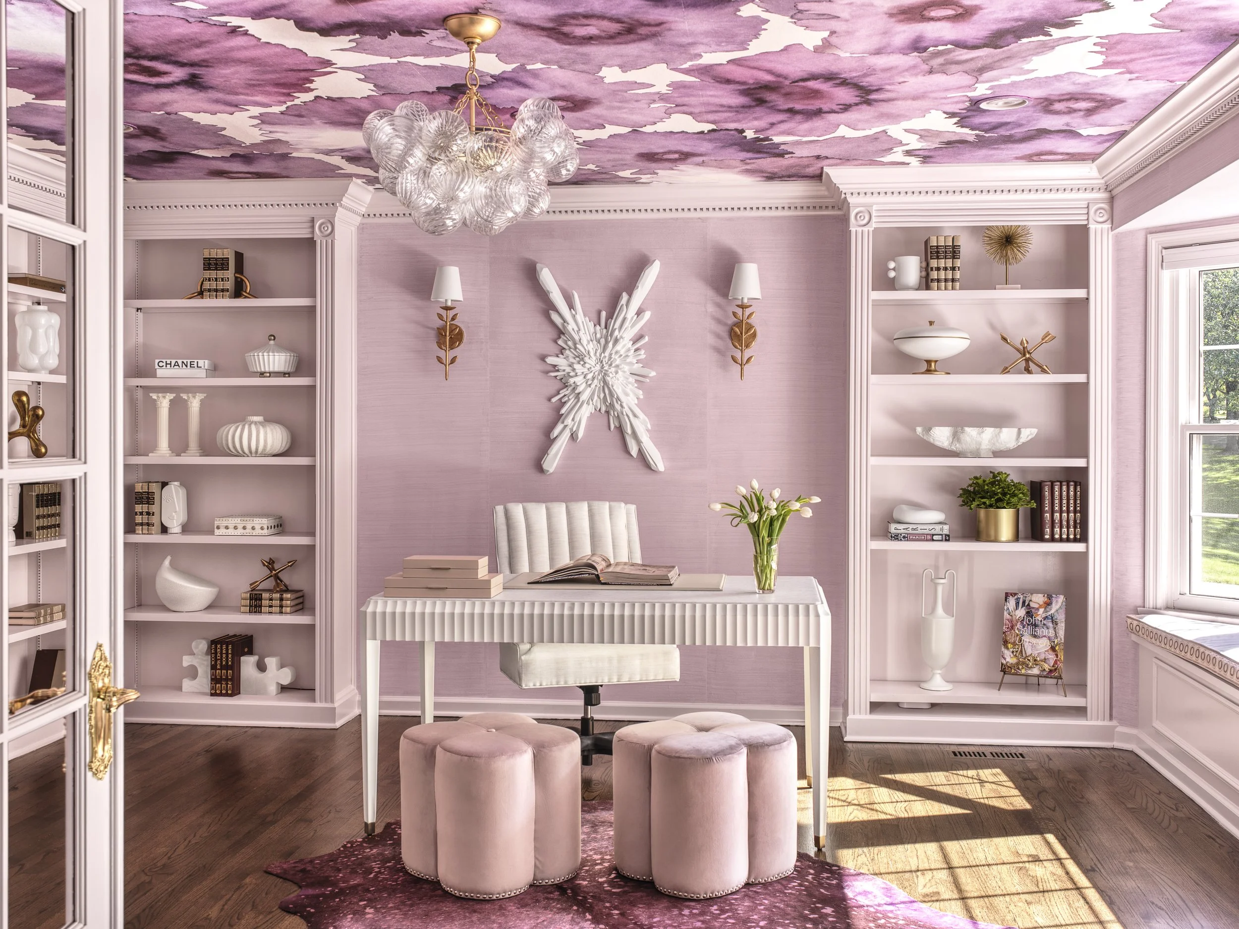 A pink and white home office with built-in bookshelves, pink tufted stools, and a white desk. The ceiling features a bold pink pattern, and an ornate chandelier hangs from the ceiling.
