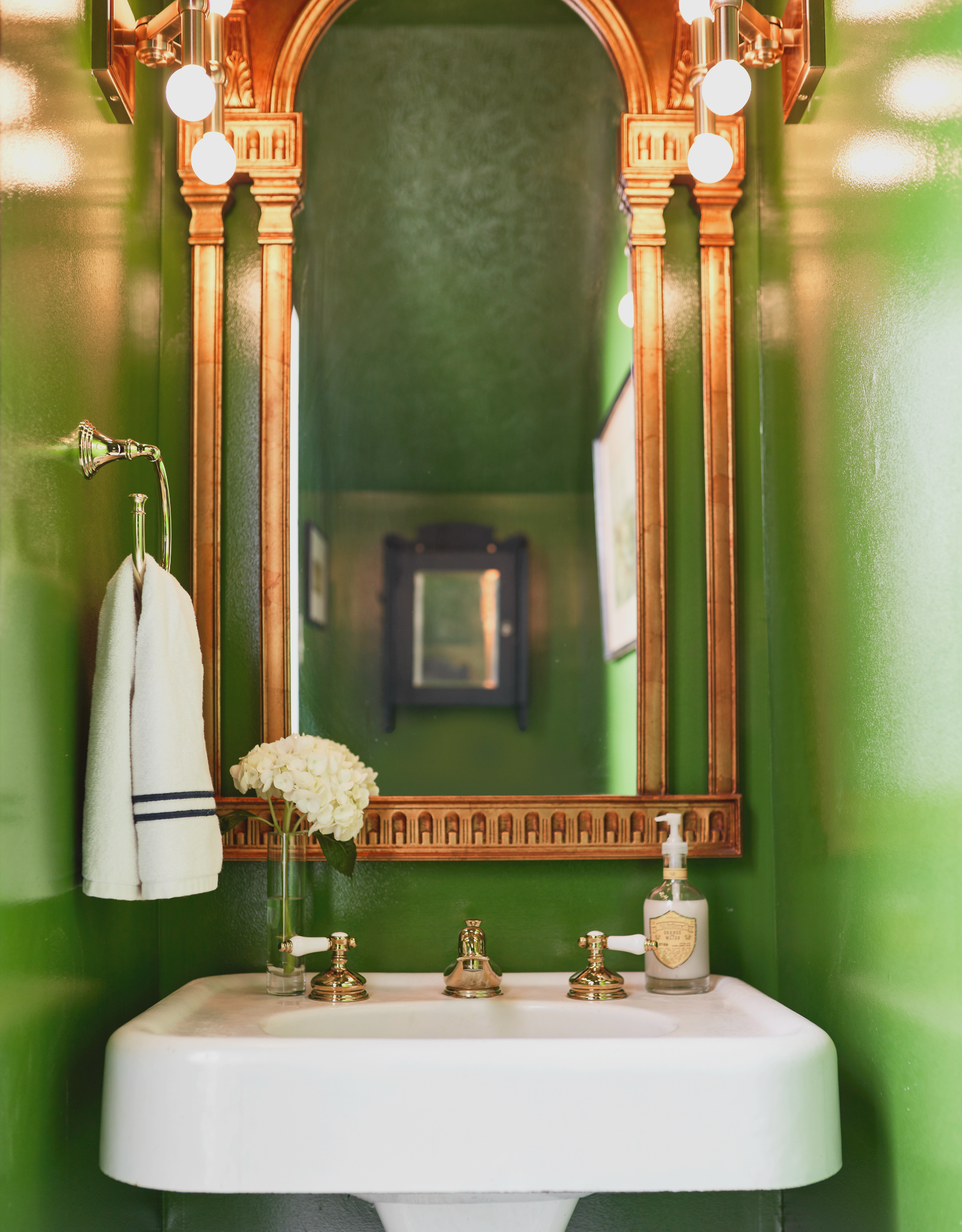 A white pedestal bathroom sink with bright green walls, a large framed mirror, and a small vase with white flowers on the sink. A towel hangs on a hook to the left, and a bottle of hand soap is on the right side of the sink.