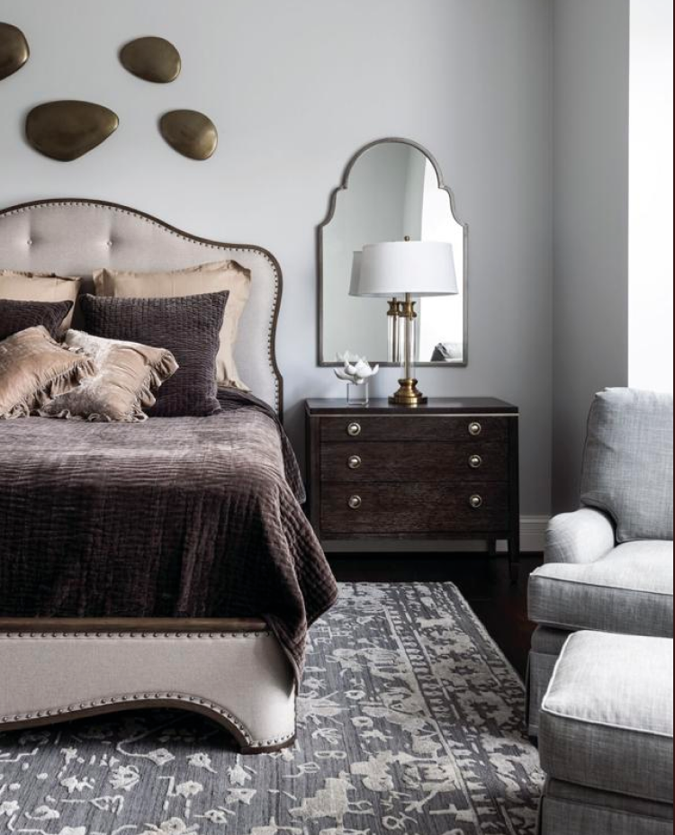 A bedroom with an upholstered bed, dark wood nightstand with a white lamp, decorative wall art, a mirror, and a gray armchair on a gray patterned rug.