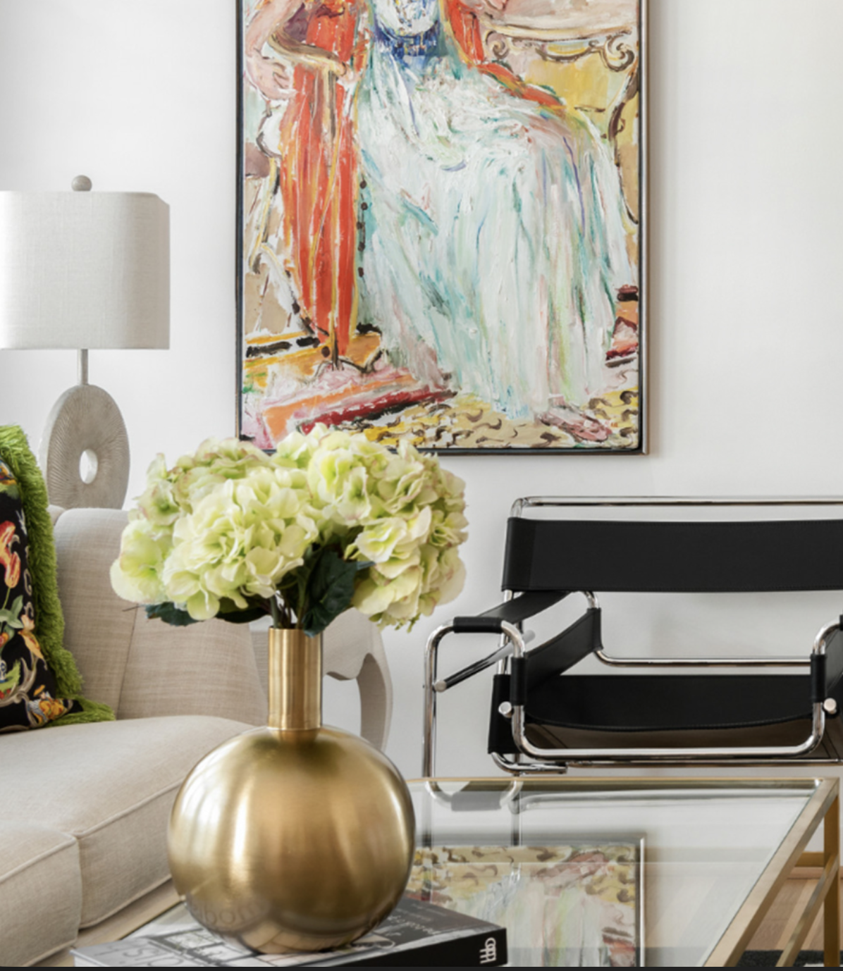 A living room with a large colorful abstract painting on the wall, a beige sofa with a dark floral pillow, a gold vase with white hydrangeas on a glass coffee table, a black modern lounge chair, and a table lamp with a circular base and a white shade.