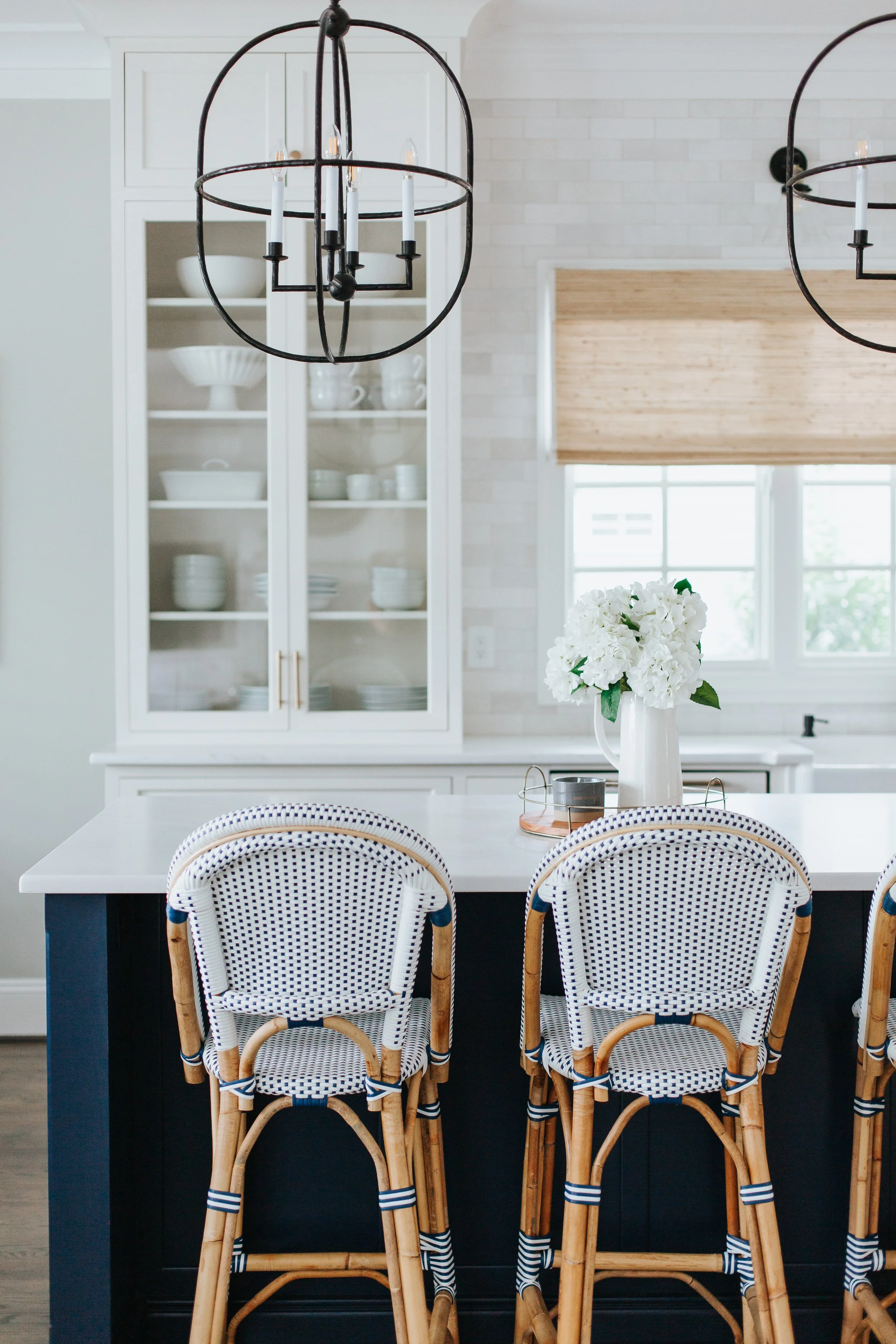 A modern kitchen with white, open shelf cabinetry and kitchen island with chairs are positioned around. The kitchen has large windows and the overall decor is light and airy.