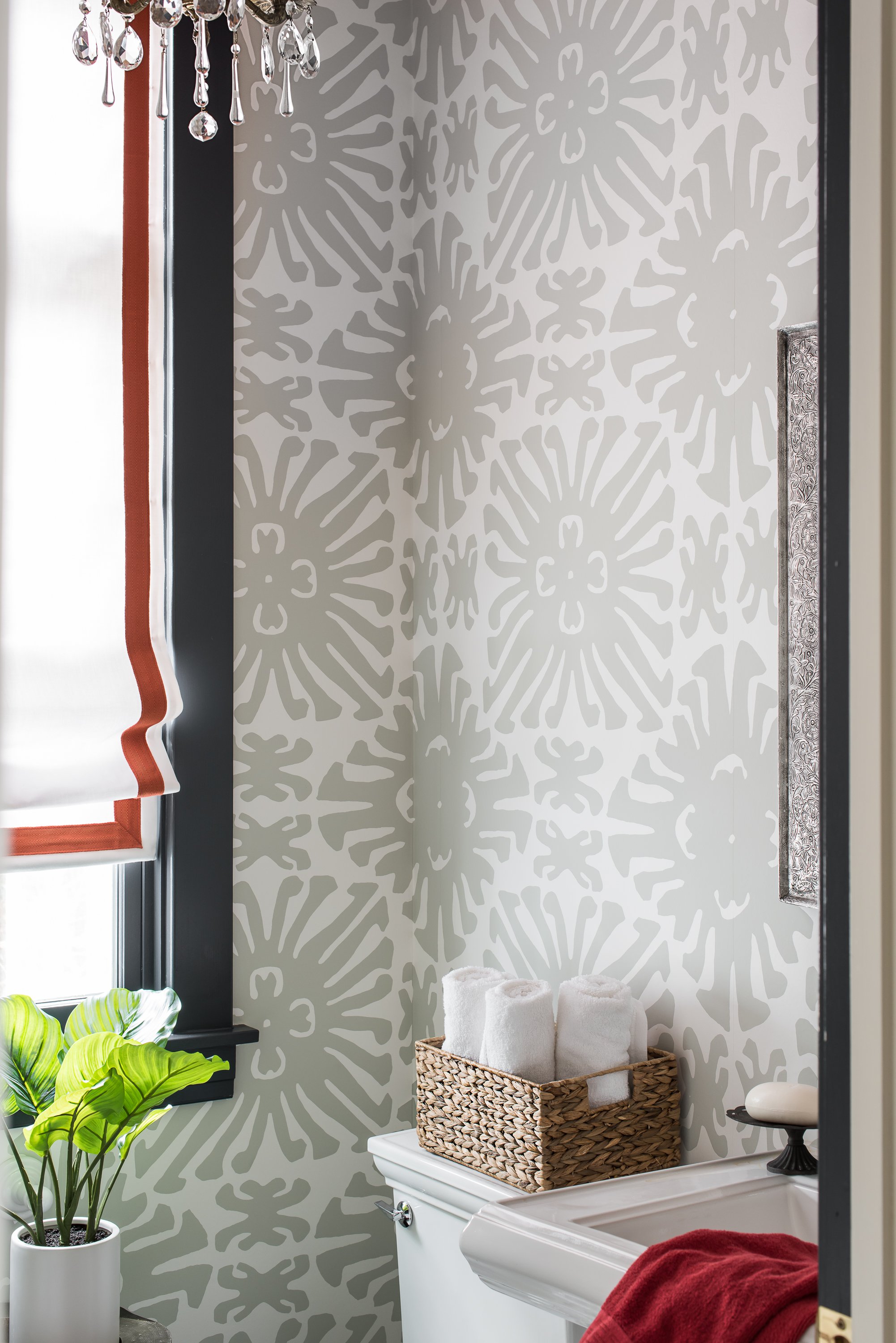A bathroom with patterned gray and white wallpaper, a basket with rolled white towels on the toilet tank, a potted green plant, a window with a black frame and a white curtain with colorful trim, and a decorative rectangular mirror.
