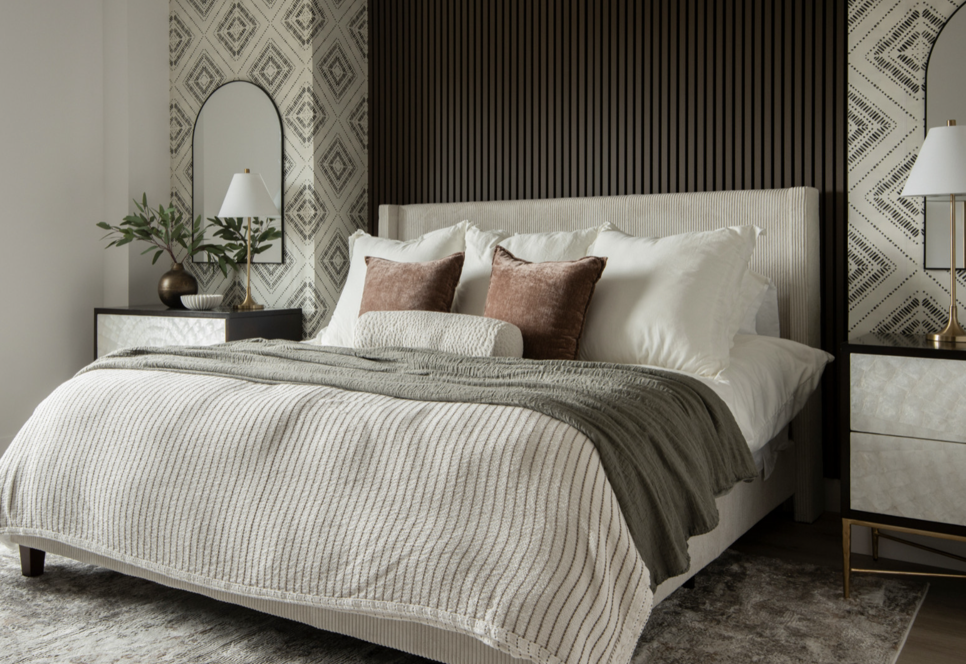 A neatly made bed with white linens, beige and brown pillows and a textured blanket, flanked by textural nightstands with pearlescent shell drawers and modern lamps, against a wall with geometric wallpaper.