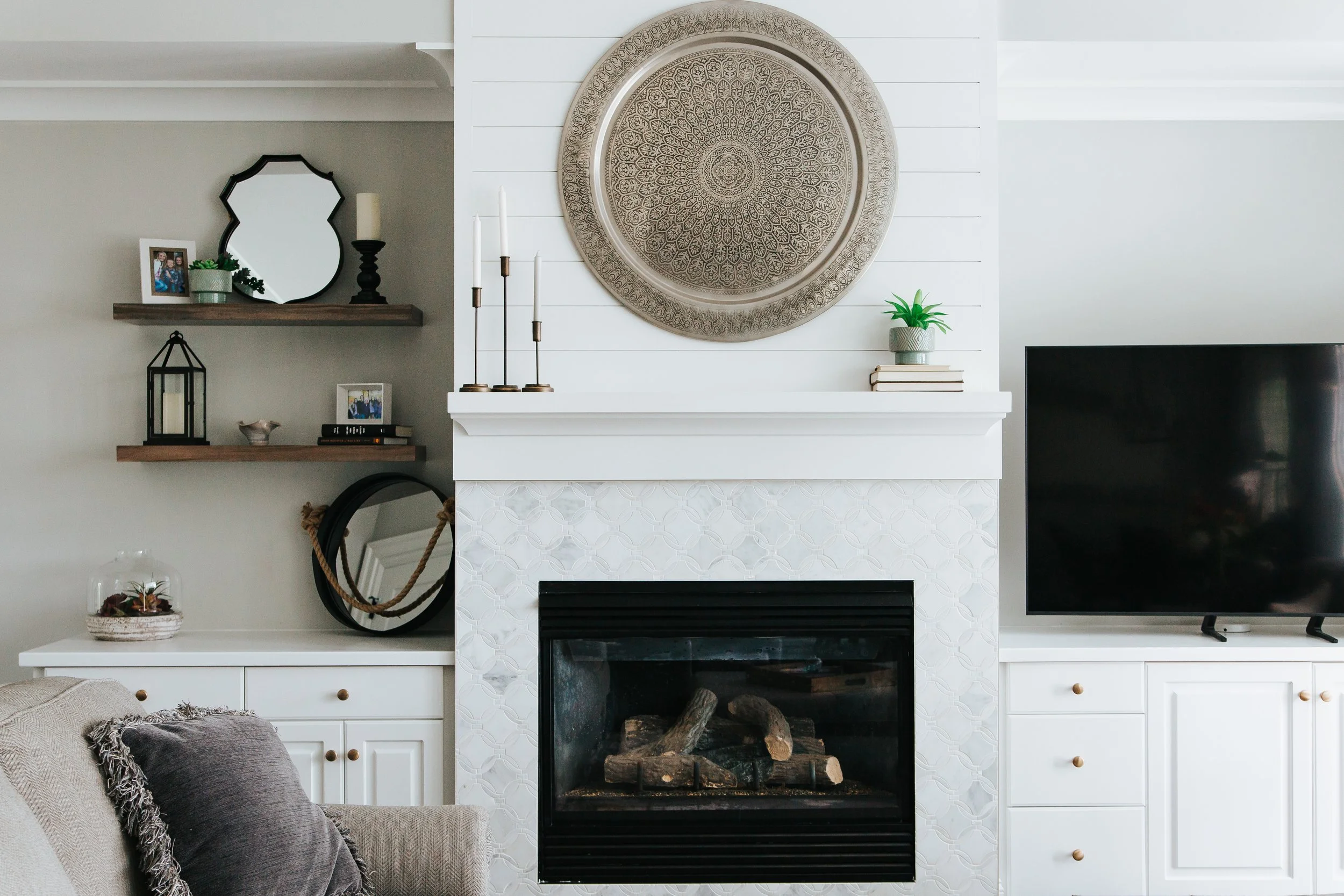 Living room with white shiplap wall, a white fireplace mantel and black accents. Shelves to the left display a mirror and other knick knacks. A large decorative circular metal wall hanging is above the fireplace.