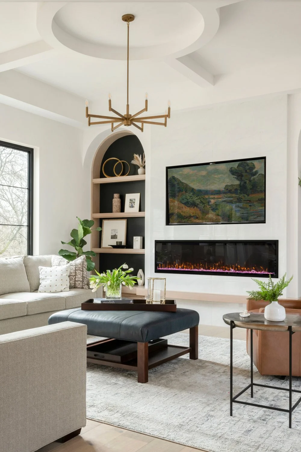 Living room with fireplace, wall-mounted TV, beige sofa, black ottoman with tray and flowers, round side table with plant, ceiling light fixture, bookshelf, and window.