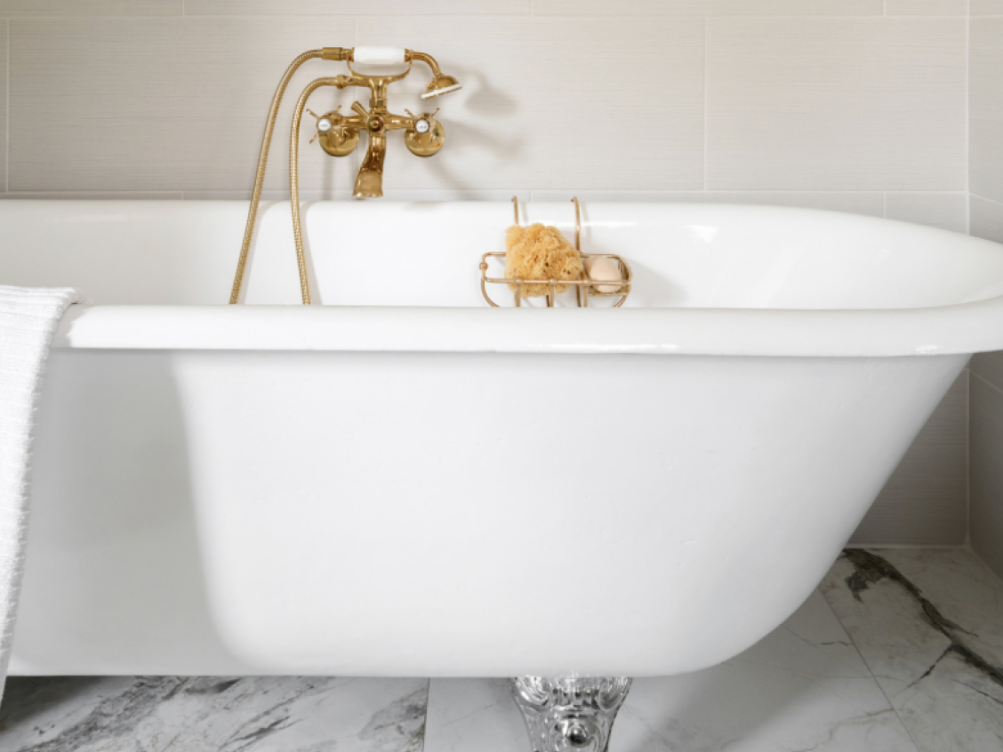 White clawfoot bathtub with gold fixtures in a bathroom with light gray tiled walls and marble floor.