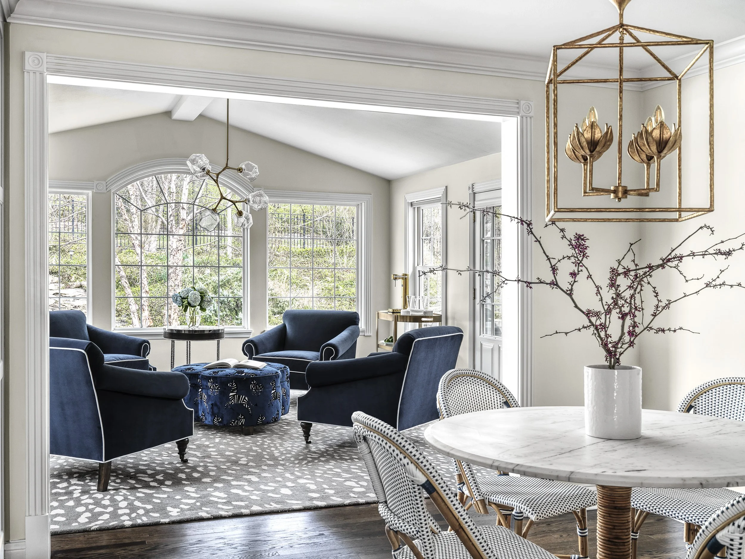 A bright living room with large windows, blue armchairs around a small table, and a chandelier with a gold edge and floral design hanging. There is a round dining table with a white marble top and wicker chairs in the foreground.
