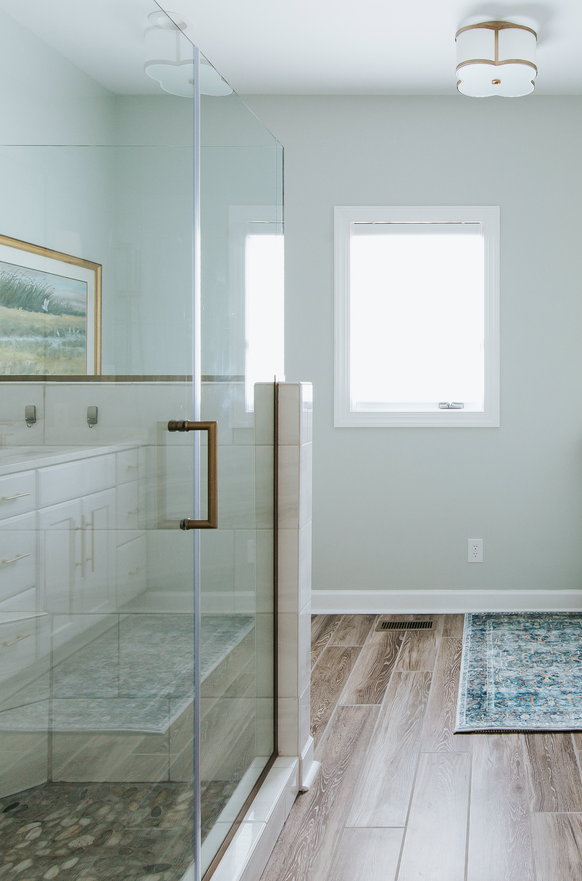 A bathroom with a glass shower with a brass handle, white cabinets, a window with white trim, and a ceiling light.