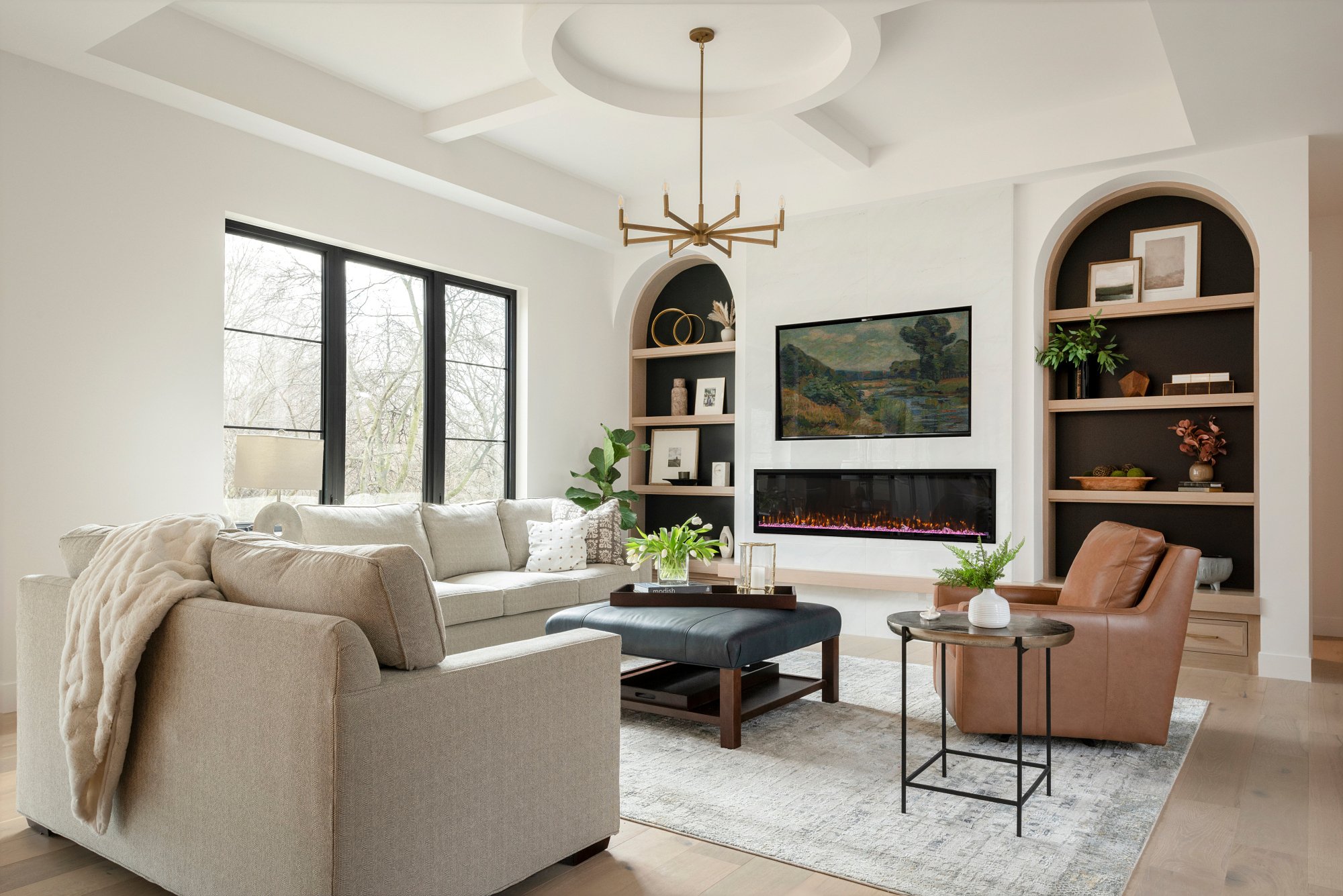 Contemporary living room with white walls, beige sectional sofa and black coffee table. Large window, modern fireplace and wall-mounted TV. Shelves with decor, minimalist chandelier, and neutral rug.