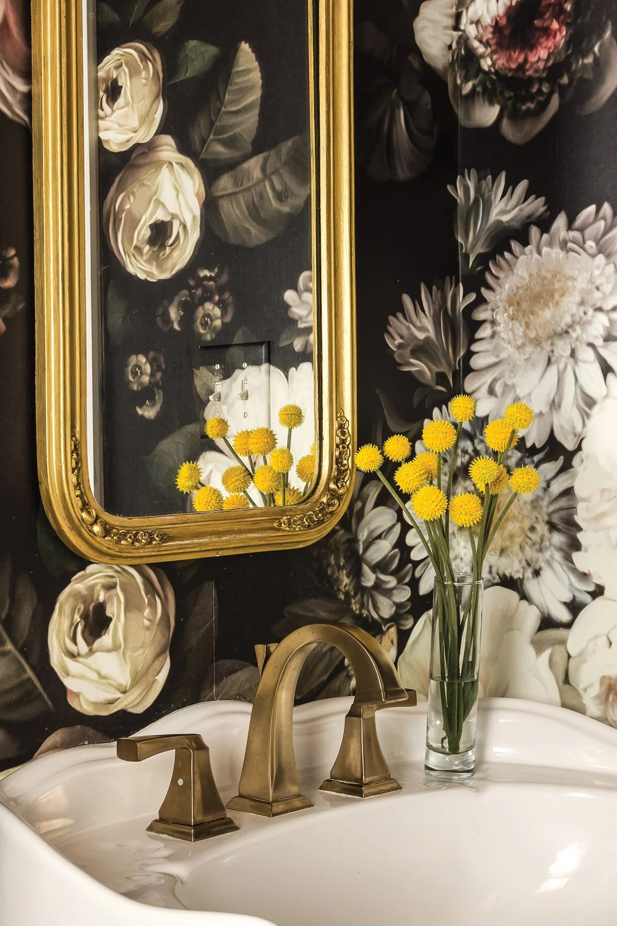 brass mirror on black floral wallpaper above cropped pedestal sink with brass faucet and yellow flowers in vase