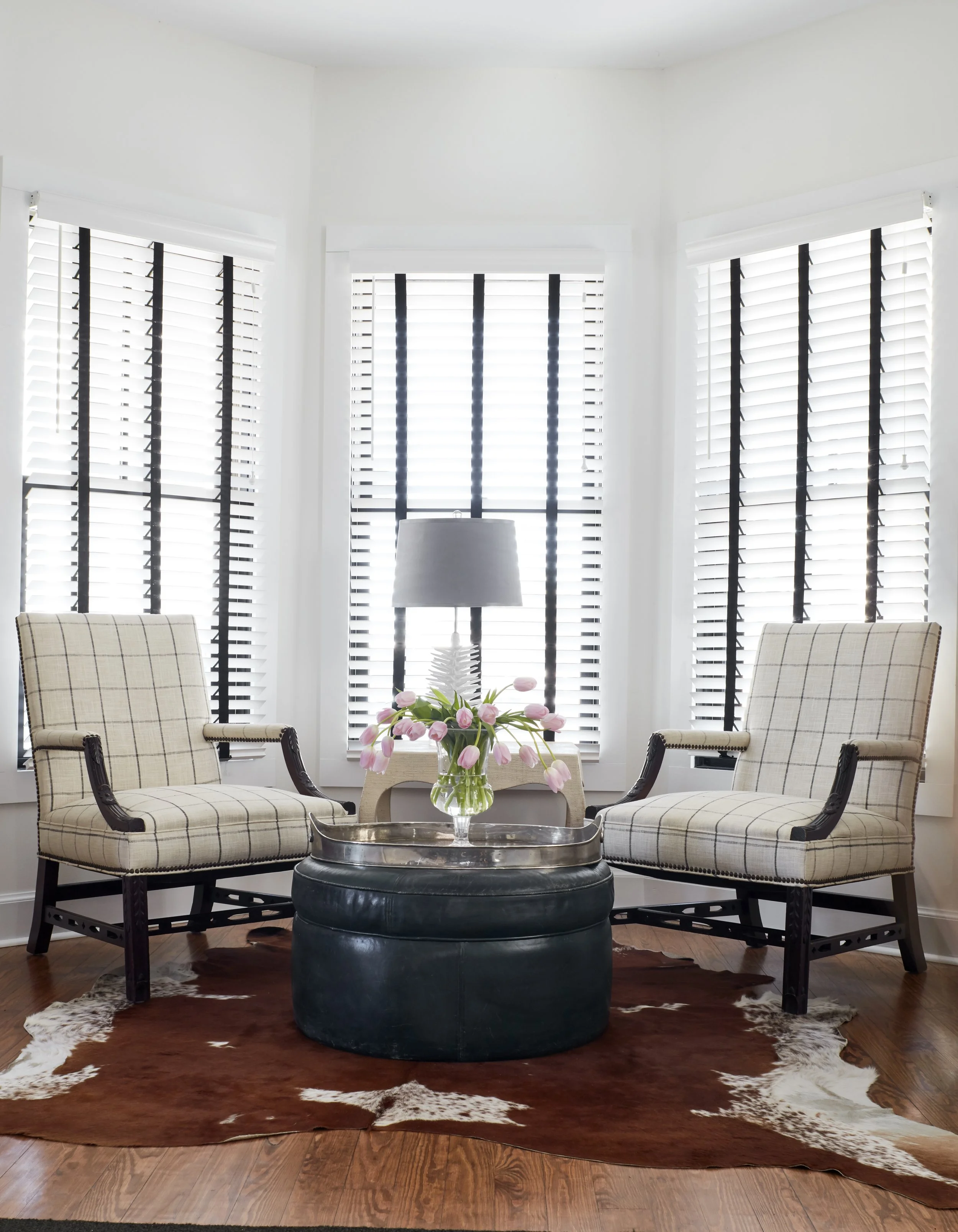 A living room with white walls and large windows with blinds, two plaid armchairs, a round black leather ottoman with a silver tray on it, a glass vase with pink tulips, a table, and a brown and white cowhide rug on wooden flooring.