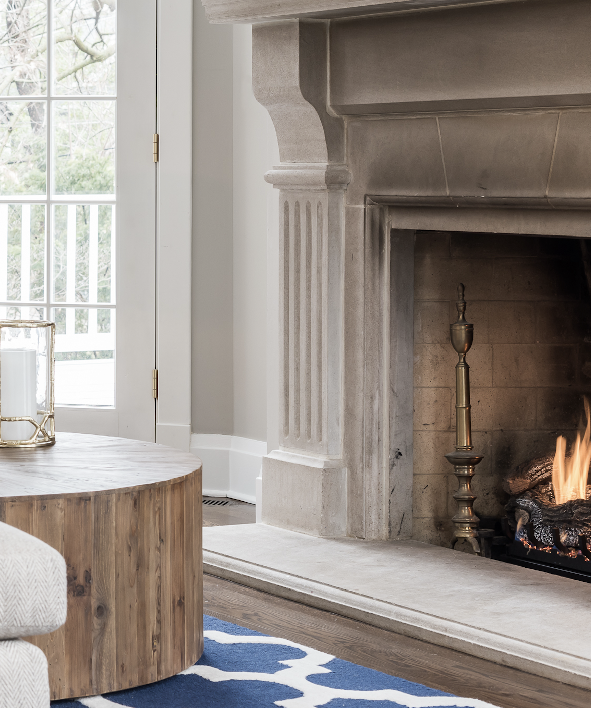 A living room corner featuring a fireplace with a brass andirons, a wooden coffee table, a blue and white patterned rug, and a window with white framing.