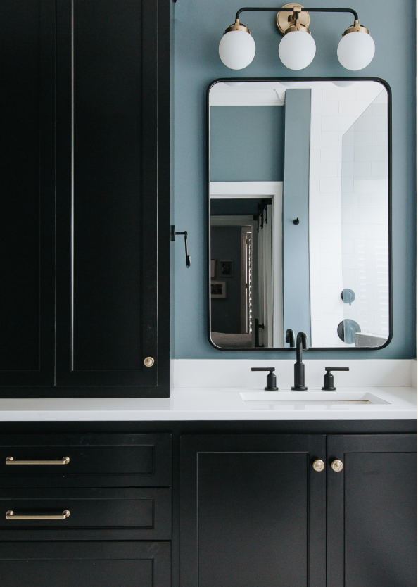 Bathroom with black cabinetry, white countertop and a large mirror. A three-light fixture with globe bulbs is above the mirror, and below, a faucet with black handles. The wall behind the mirror is bold blue.
