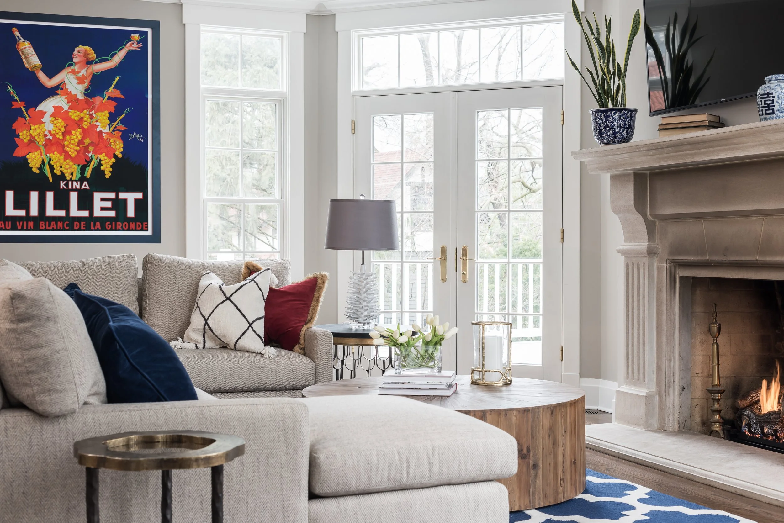 A cozy living room with a beige sectional sofa decorated with pillows, a fireplace with a mantel, a large plant in a blue and white pot, a wall-mounted television, a lamp on a side table, a pile of books, a glass vase with white flowers, and a large framed vintage-style poster on the wall.