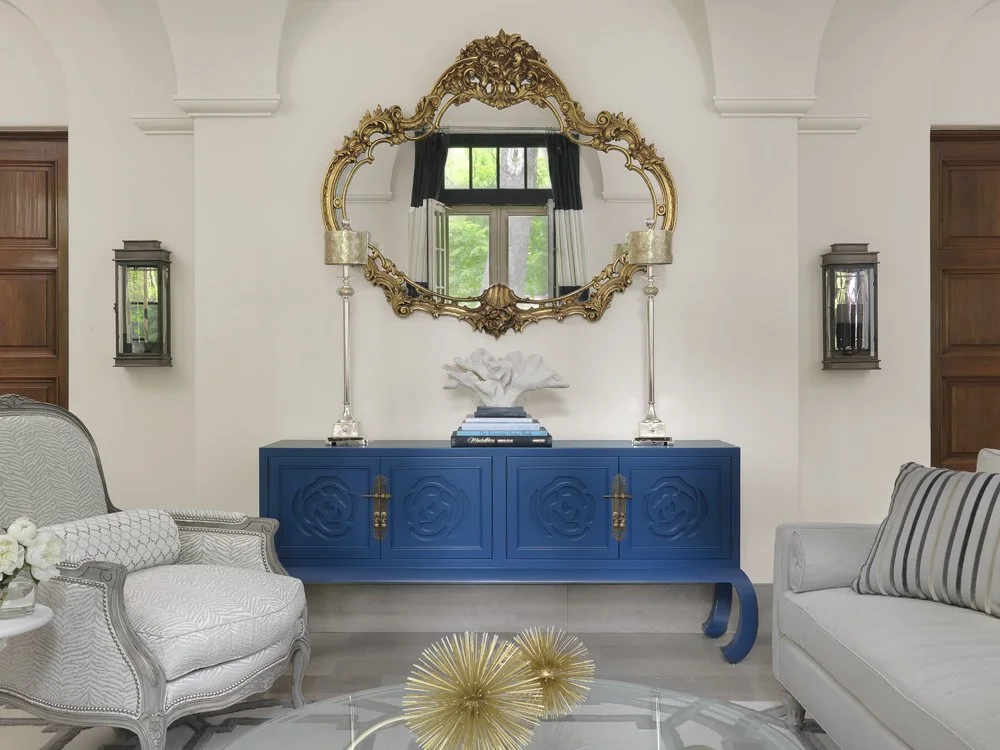 Interior of a living room with a blue sideboard and large ornate gold-framed mirror above. Two tall silver lamps flank the mirror. On the left, a patterned armchair, and on the right, a sofa with striped pillows. A glass table with gold decorative objects is in the foreground.