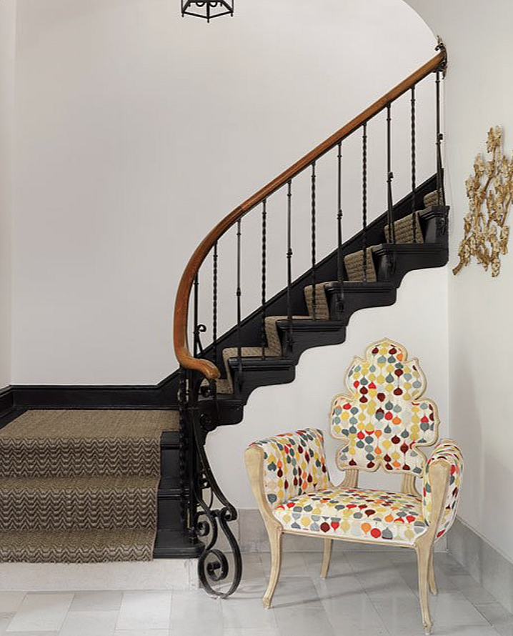 Interior of a house with a curved staircase featuring a wooden handrail and black wrought iron, an upholstered armchair with a colorful polka dot pattern, and a decorative wall art piece.
