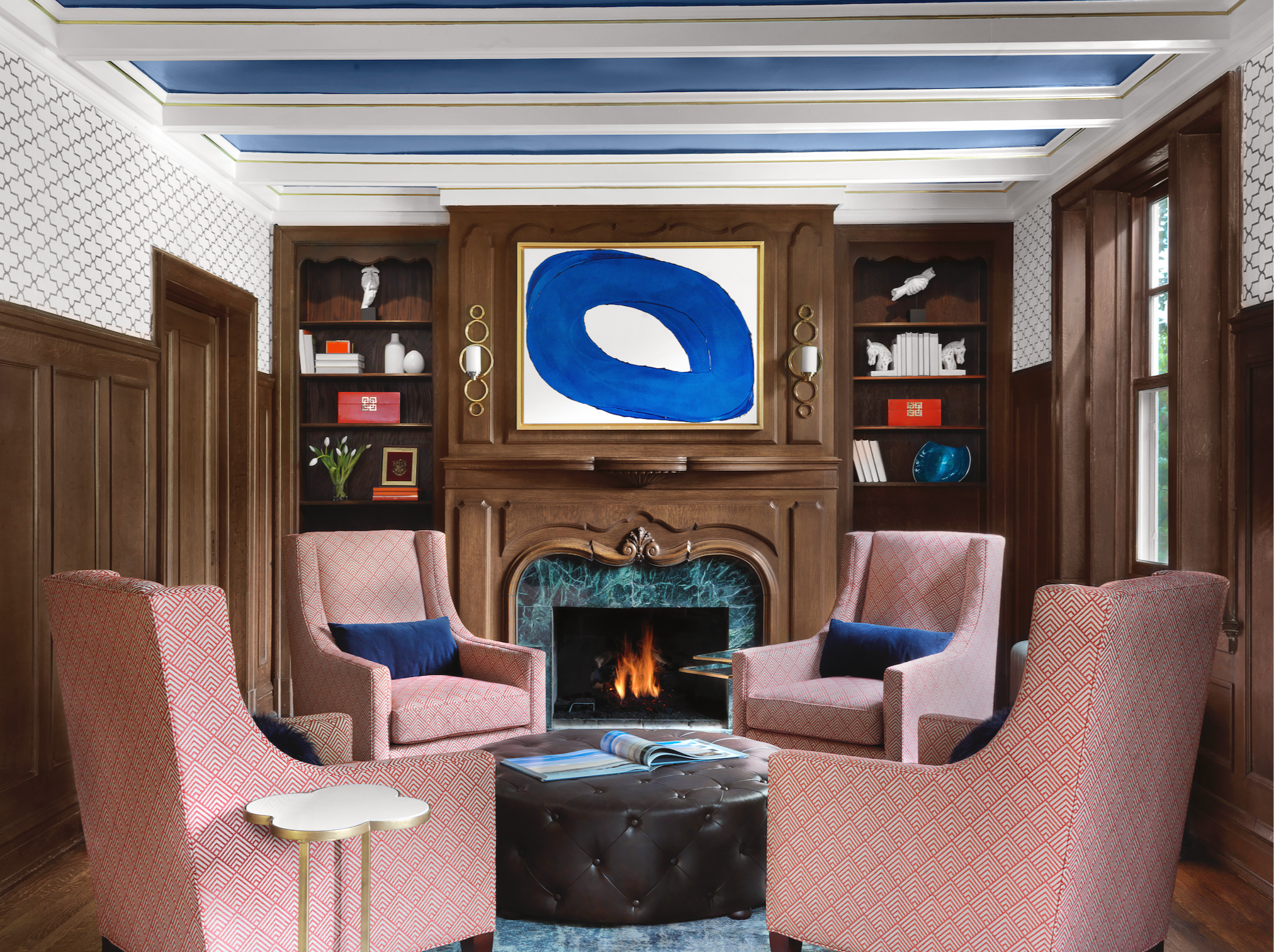 Living room with fireplace and blue contemporary artwork, patterned armchairs, blue cushions, a coffee table with books, wood-paneled walls, built-in shelves with decorative objects, and curtains on windows.