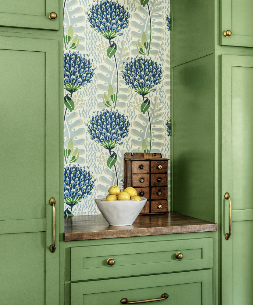 A kitchen corner with green cabinetry, a wood countertop, a white bowl filled with lemons, and wallpaper with large blue and green floral pattern.