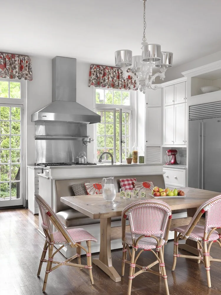 A kitchen with white cabinets, a stainless steel range hood, a wooden dining table with six chairs, a chandelier, and large windows with floral valances.