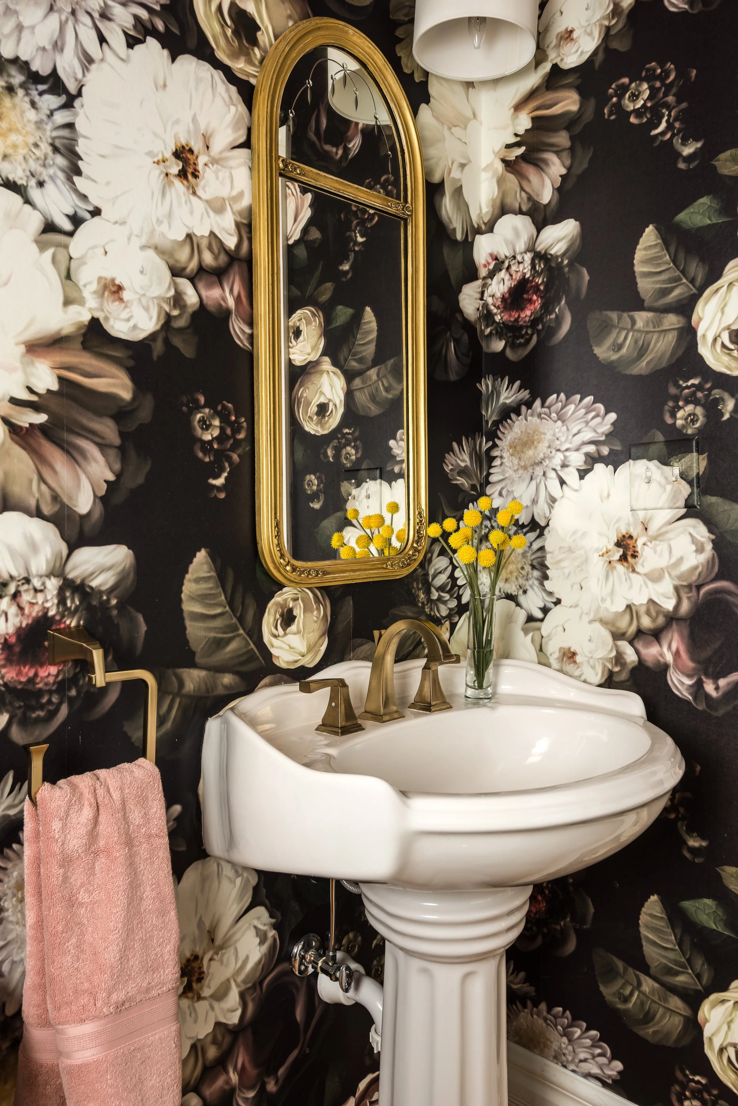 A white pedestal sink with gold fixtures and a decorative gold-framed mirror above. The wall has black wallpaper with large white and pink peony flowers. A pink towel hangs on a gold towel holder to the left of the sink, and a clear glass vase with yellow flowers is placed on the sink.
