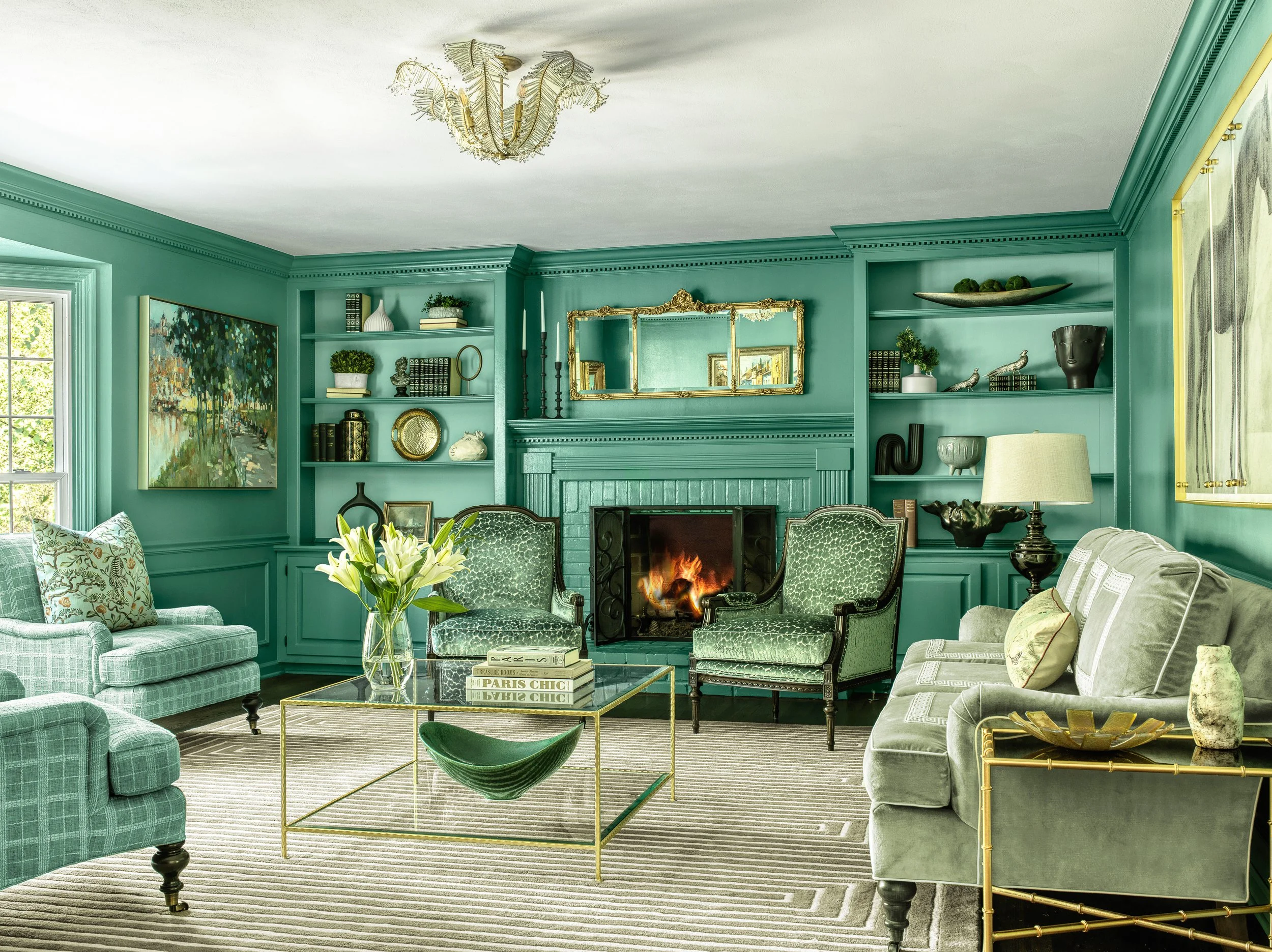 A living room with peacock green walls and built-in shelves with decorative objects, a fireplace, patterned armchairs, a sofa with patterned pillows, a glass coffee table with a vase of flowers, and a striped rug. A chandelier hangs from the ceiling.