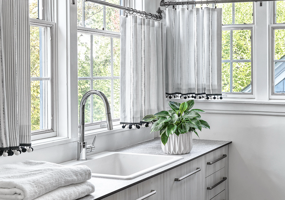 A bright kitchen window with black and white patterned curtains, a white sink, a chrome faucet, a potted green plant on the countertop, and folded white towels.