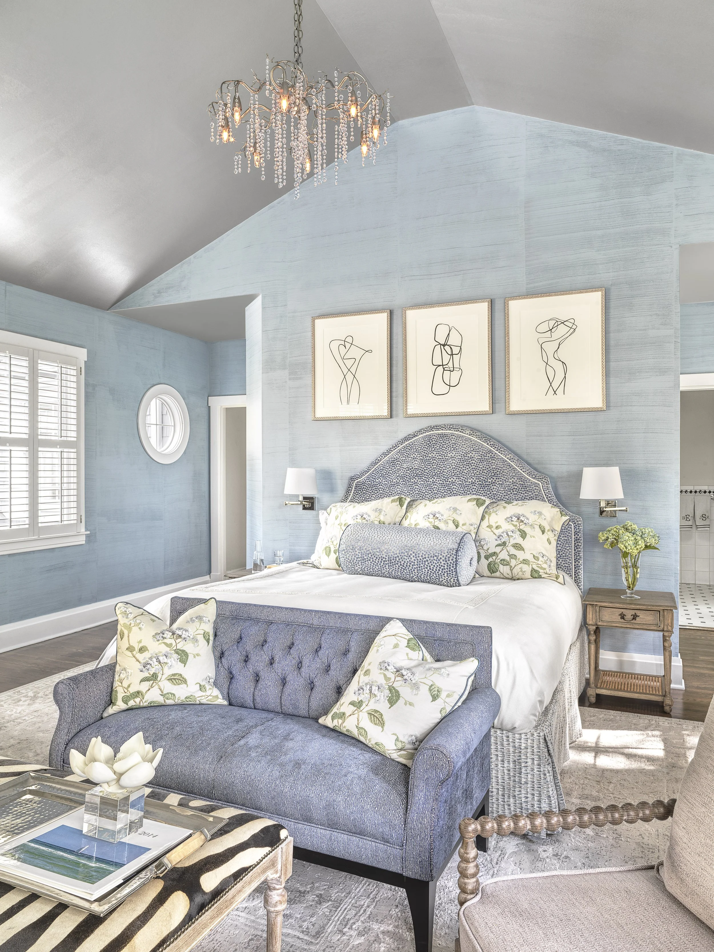 A bedroom with blue wallpaper, bed with floral and blue pillows, a wooden side table with a vase of flowers, a blue upholstered sofa at the foot of the bed, and a chandelier.