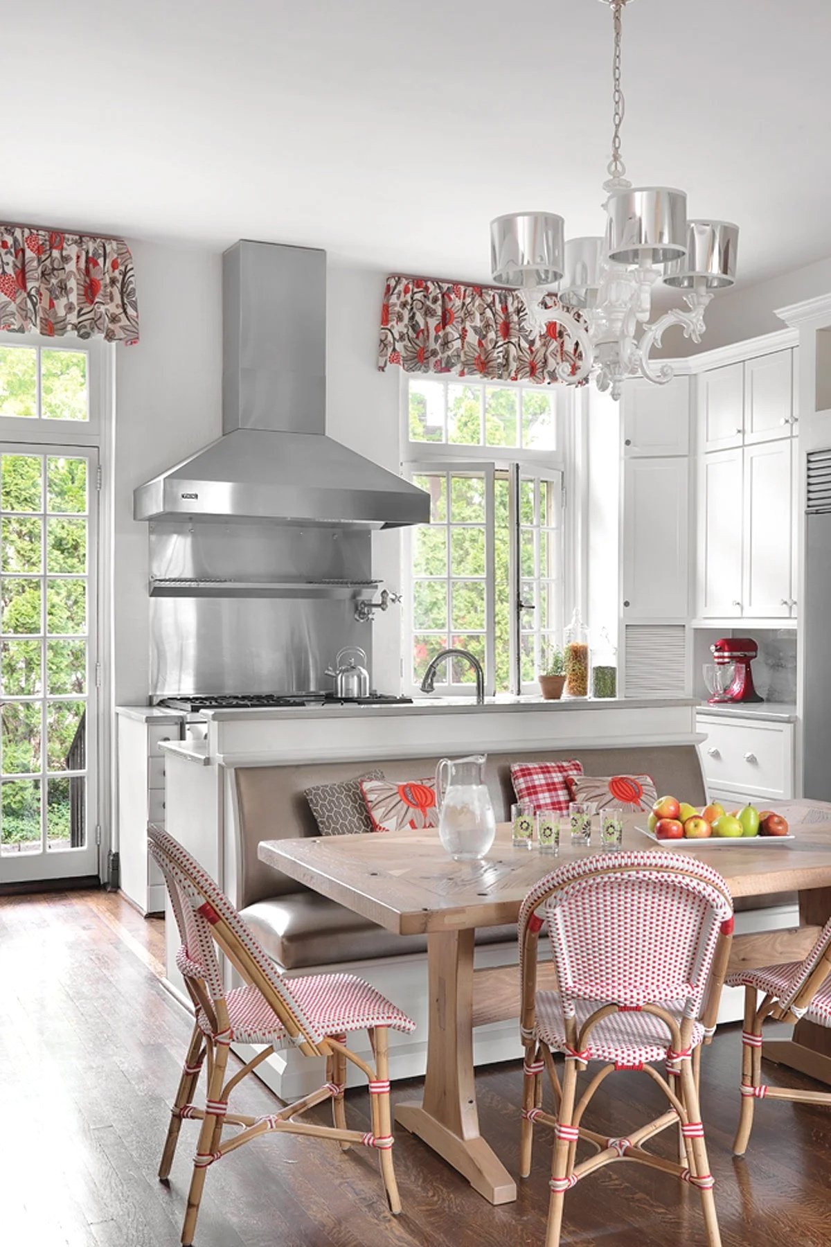 A bright kitchen with white cabinetry, a wooden dining table, and red and white patterned chairs. There are windows with floral curtains, a stainless steel range hood, and a chandelier hanging from the ceiling.