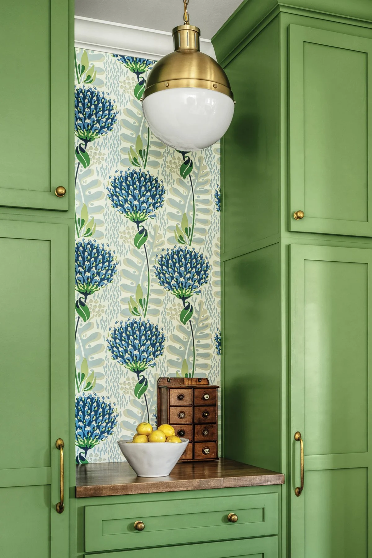 A kitchen featuring bright green cabinets, a brass and glass globe pendant light, floral wallpaper with blue and green flowers, and brass handles and knobs.