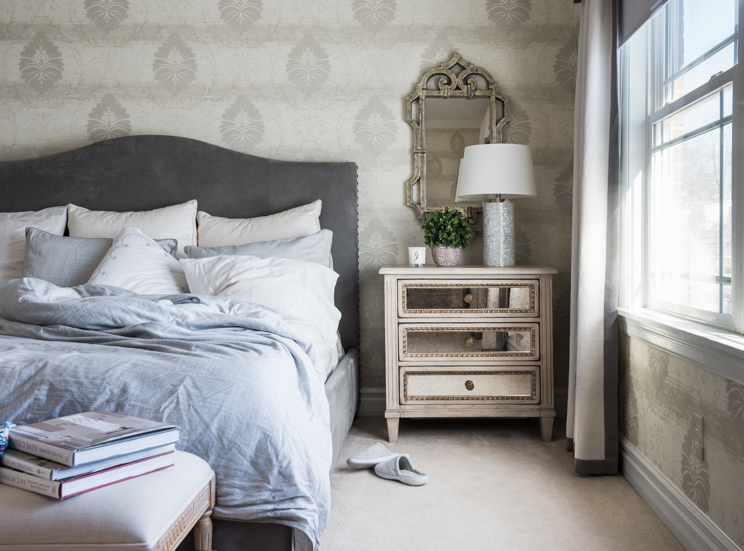 A cozy bedroom with a bed covered in white and gray bedding, a white nightstand with three drawers, a decorative framed mirror, a white table lamp, a small potted green plant, and a window with white curtains.