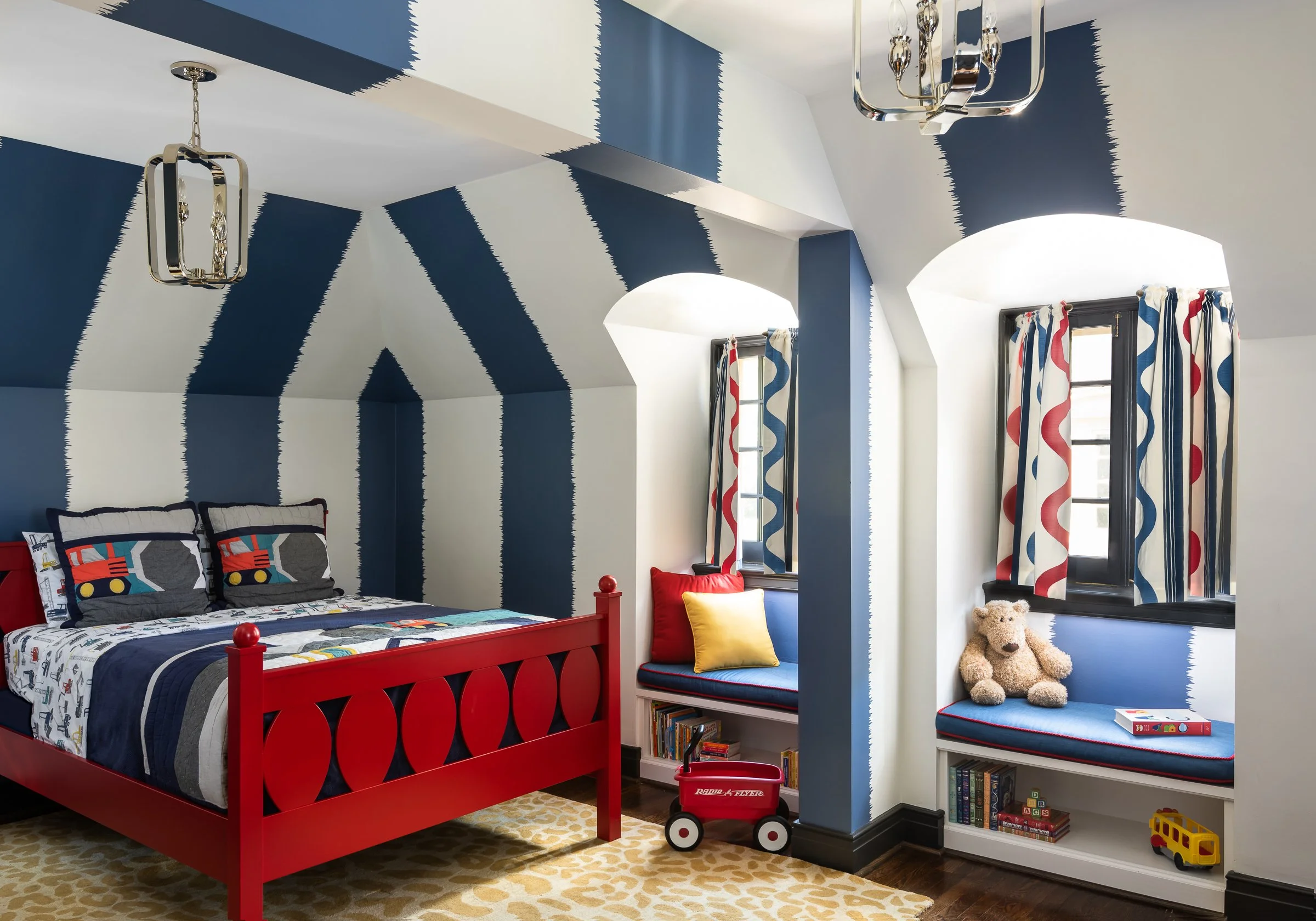 A children's bedroom with a red bedframe, blue and white striped wall and ceiling, windows with red, blue, and white curtains, and a teddy bear on a blue window seat.
