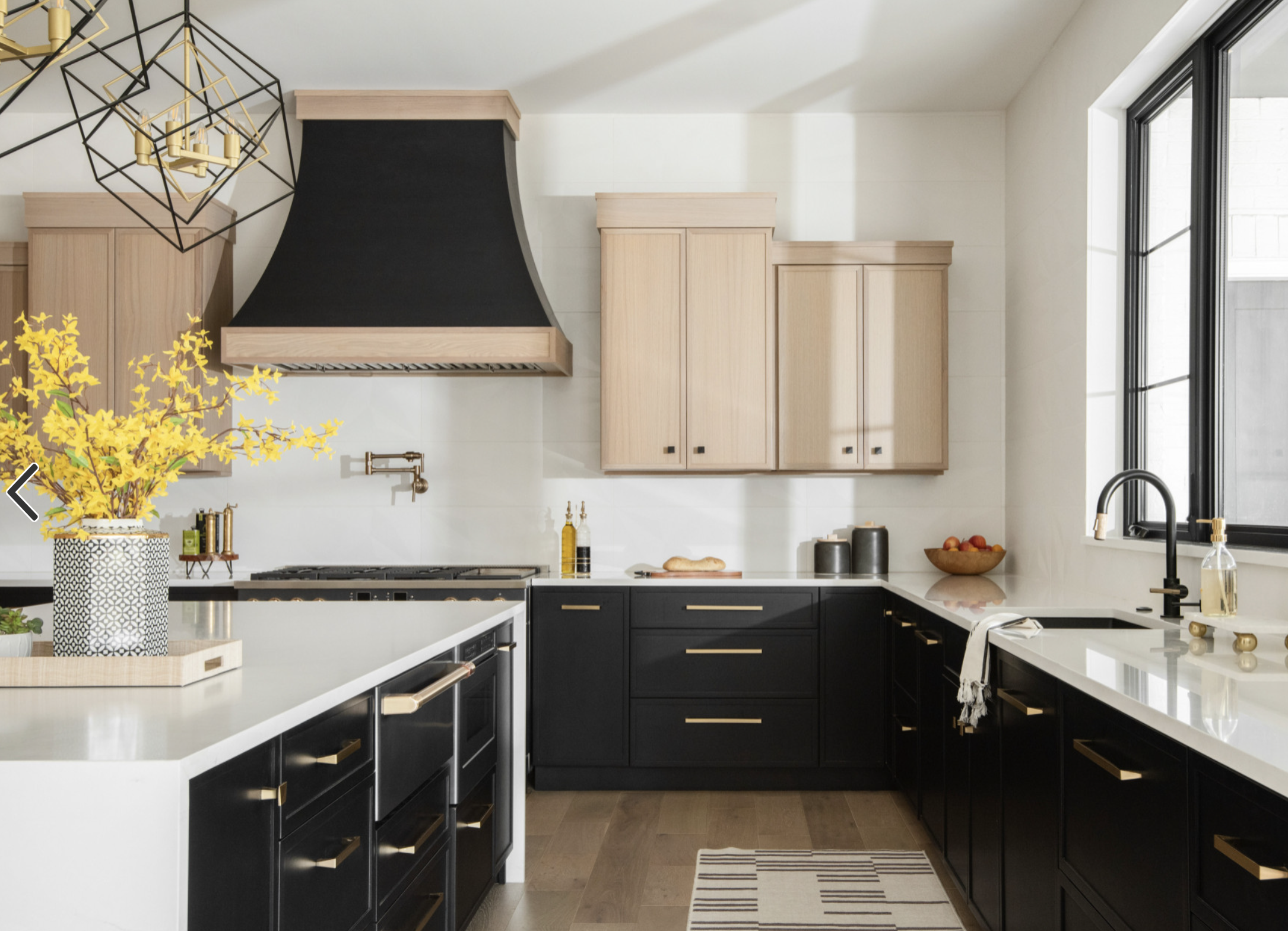 Modern kitchen with black and natural wood cabinets, white countertops, gold hardware, a black range hood and large window with black frame.
