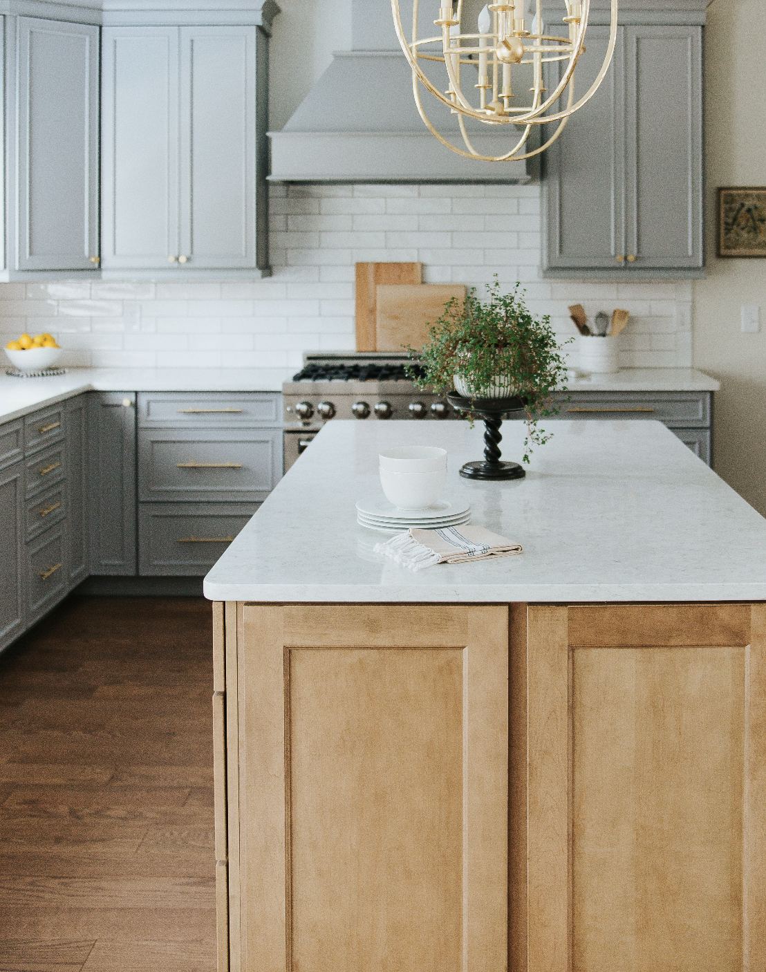 A modern kitchen with a beige island in the foreground, gray cabinets, a white marble countertop, a stainless steel stove, a white backsplash, and a chandelier hanging above the island. Decor includes a potted plant, a bowl of lemons, and ceramic bowls.