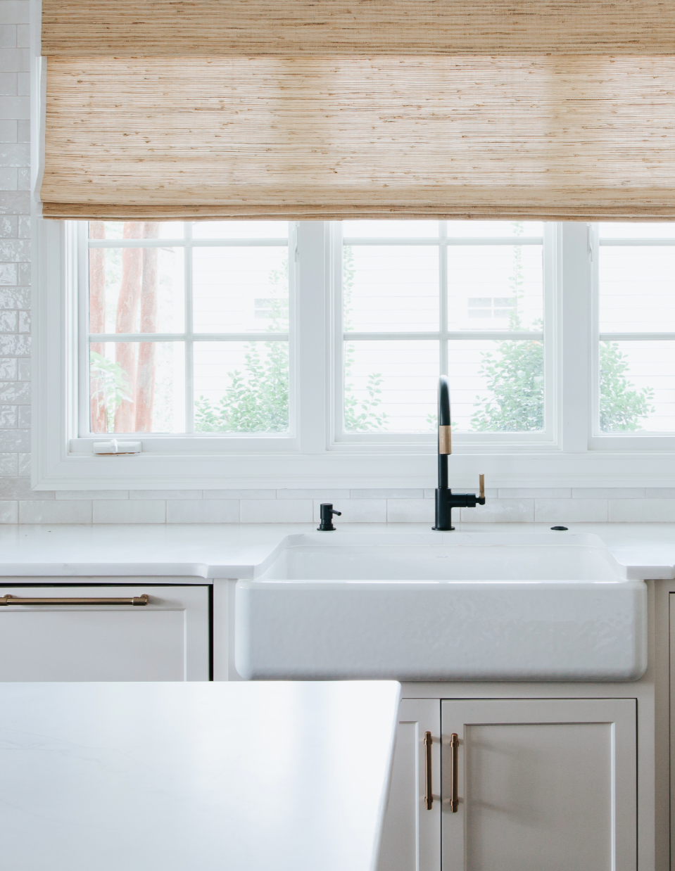 Modern kitchen with light beige cabinetry, white sink and countertops, and a woven curtain above the sink looking out onto greenery.
