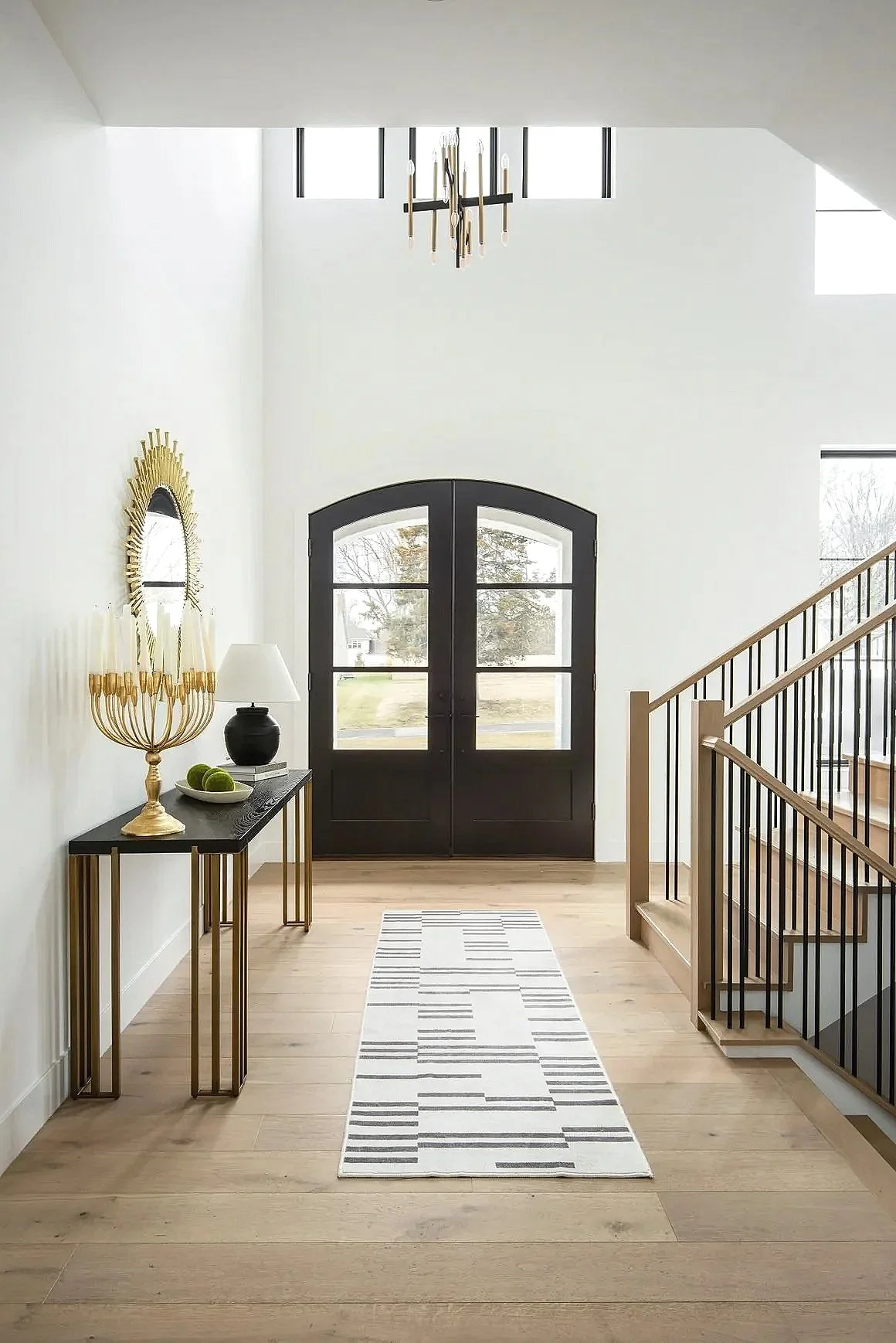 Entryway with arched black doors, staircase and a black console table. There is a modern chandelier, a white and gray striped rug, and large windows letting in natural light.