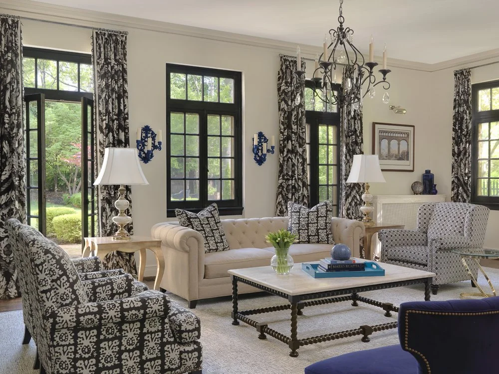 Living room with black and white patterned curtains, black-framed windows, a beige tufted sofa with black and white pillows, patterned armchairs, white tables with lamps, a central coffee table with decorative objects, and a chandelier hanging from the ceiling.