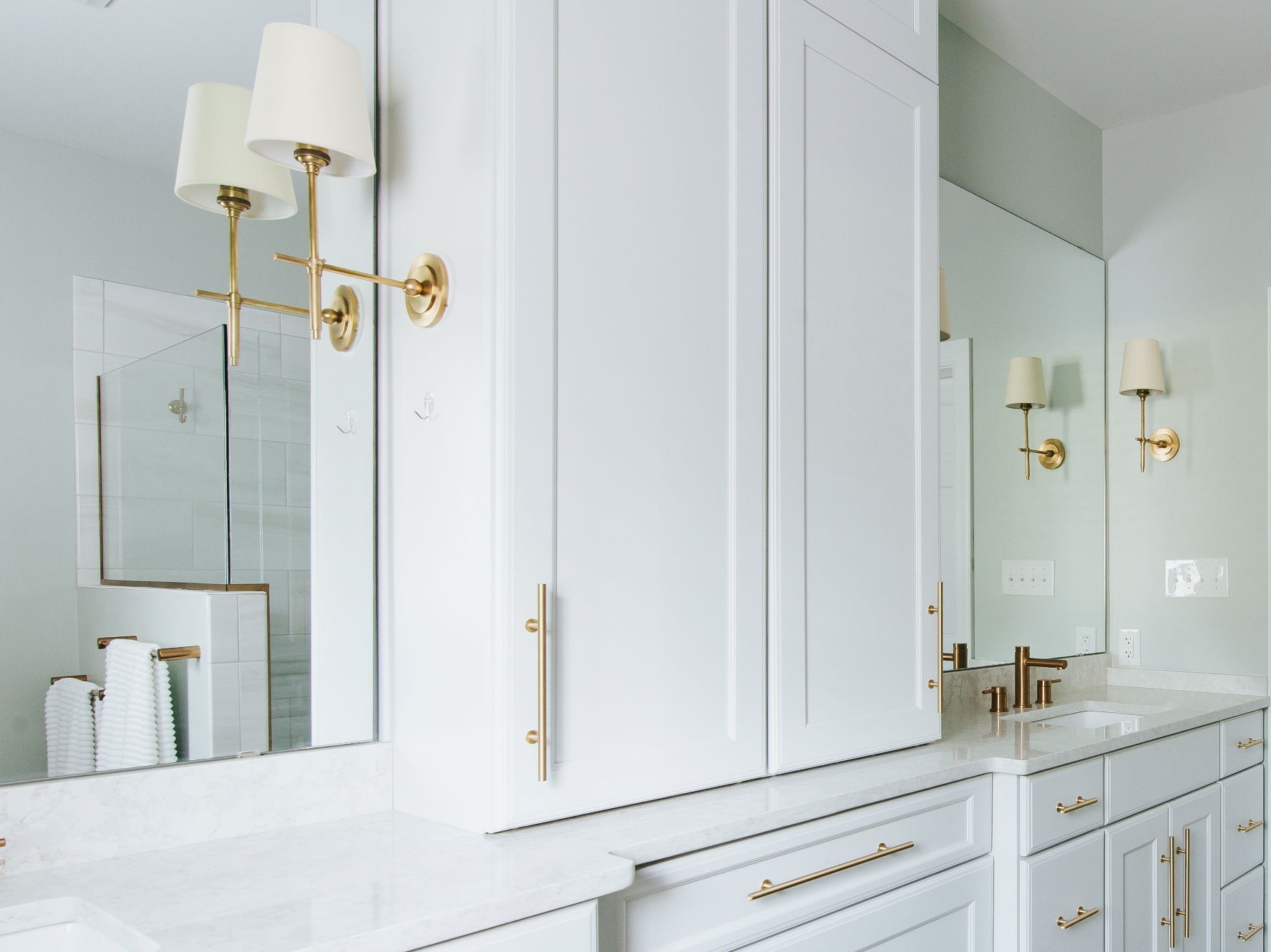 A modern bathroom with white cabinetry, gold fixtures, and large mirrors. There are white countertops and wall-mounted gold lamps with white shades.
