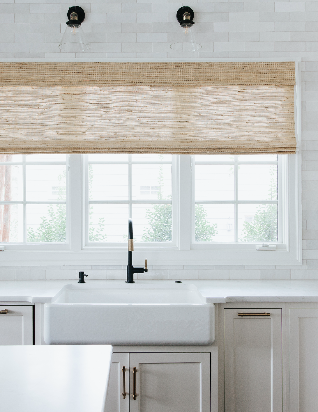 Kitchen window with white frame and woven shade above matte black faucet, white farmhouse sink and white cabinetry. Two light fixtures feature glass shades.