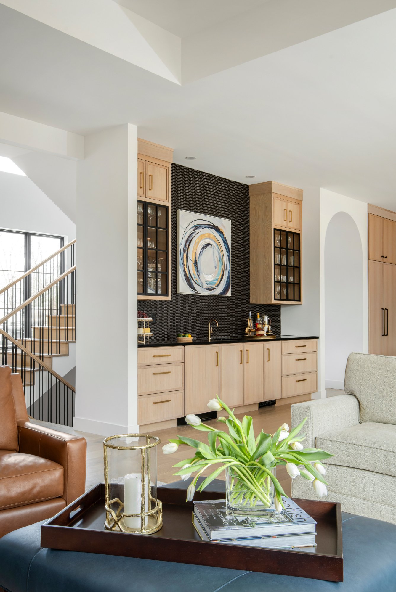 Living room and bar with light wood cabinetry, black backsplash and modern artwork. Centered is a coffee table with a tray holding candles, next to a beige armchair and a brown leather sofa.