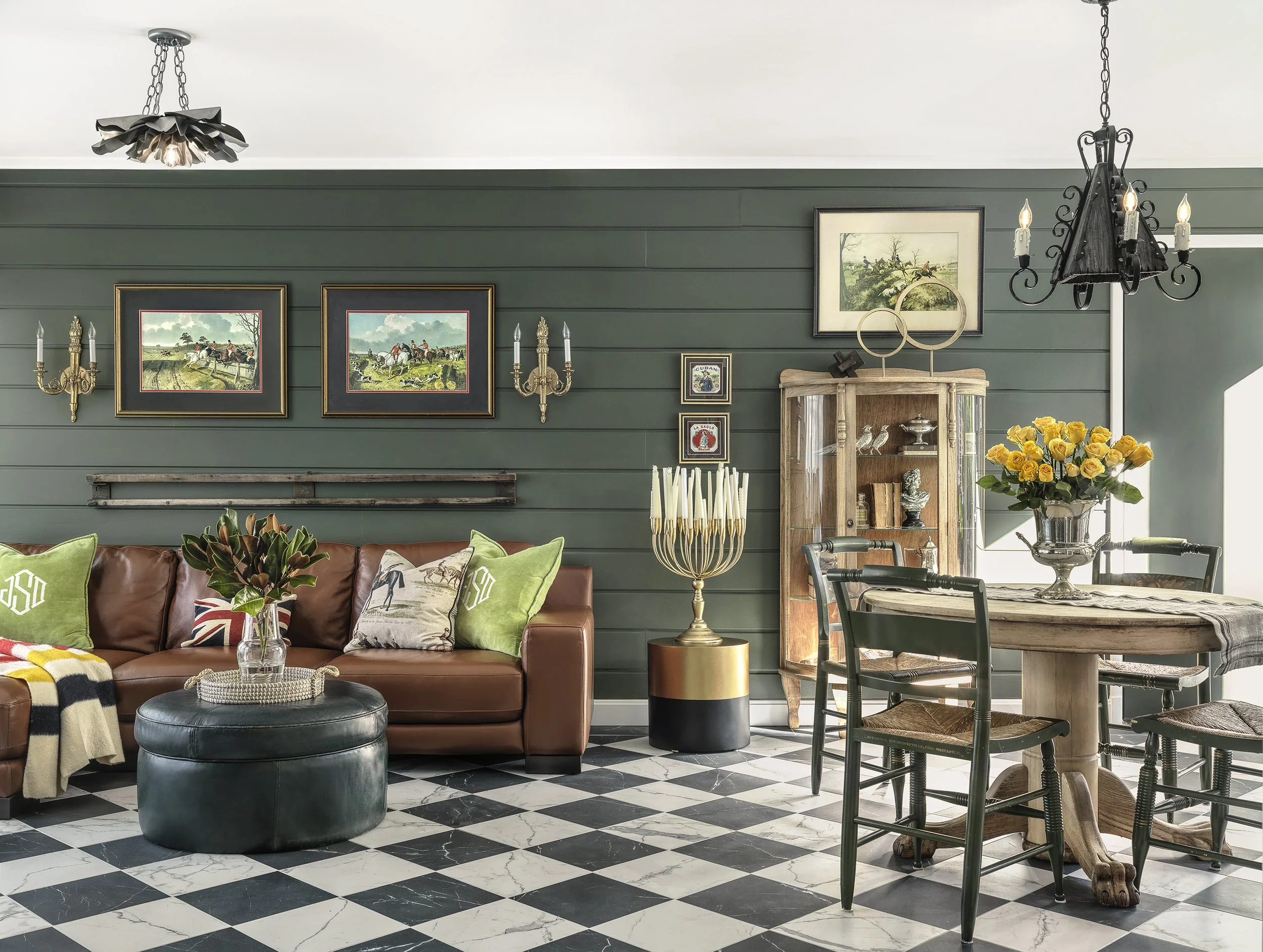 Living room with green shiplap walls, black and white checkered floor, framed paintings, antique wall sconces, chandelier, and a leather sofa with pillows.
