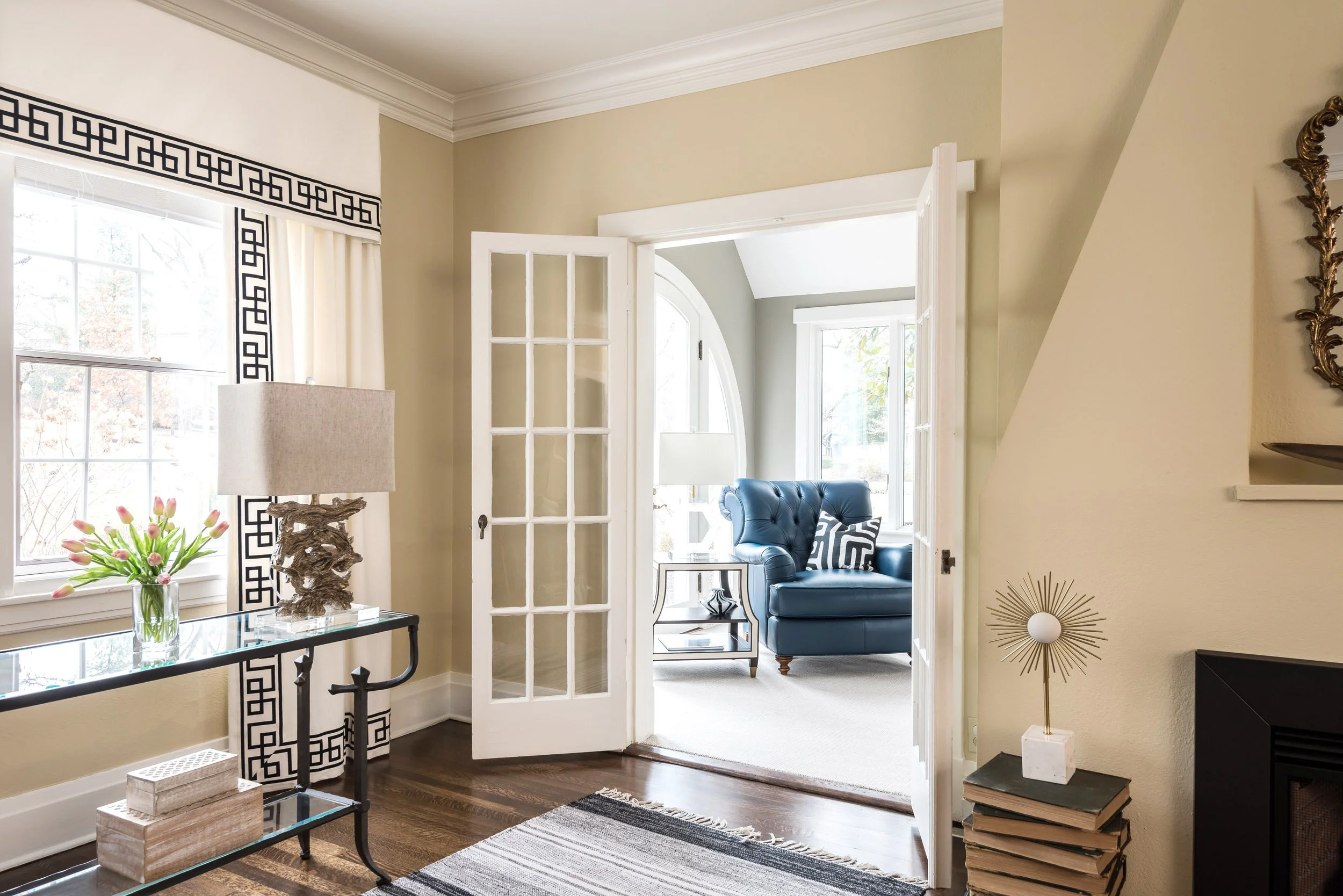 Living room with a door opening to sunroom. Inside the sunroom, a blue armchair, a side table, and large windows during daytime.