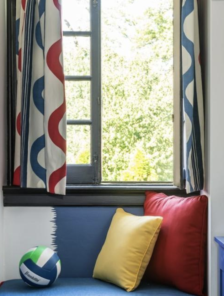 A window with colorful, patterned curtains in red, blue, and white. Outside, green trees are visible. Below the window, there's a blue window seat with a red pillow, a yellow pillow, and a multicolored volleyball.