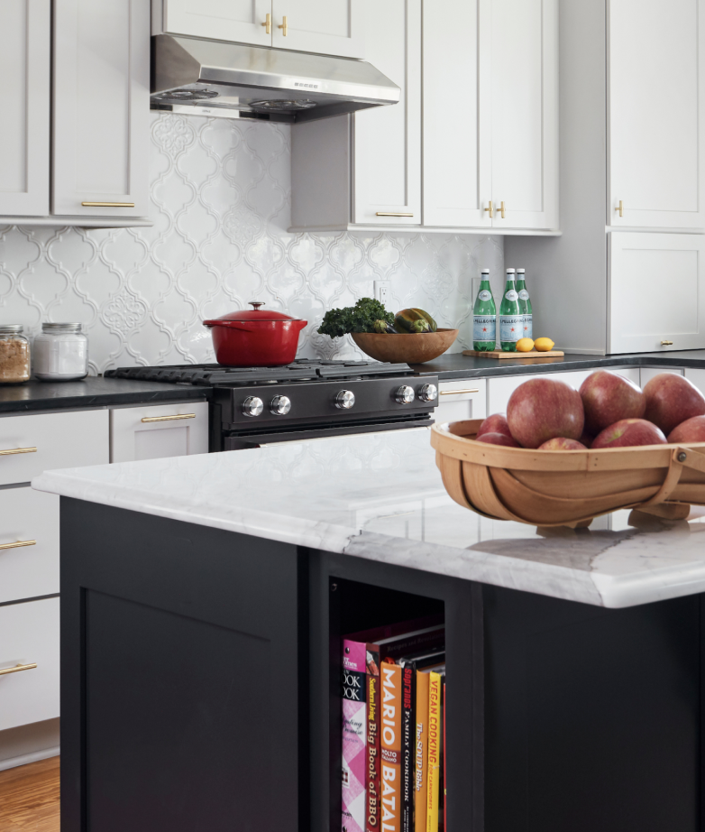 Modern kitchen with white cabinets, a black stove with a red pot, a wooden bowl with greens and vegetables, bottled water, lemons, jars of ingredients, and a black island with books and a bowl of apples on a marble countertop.