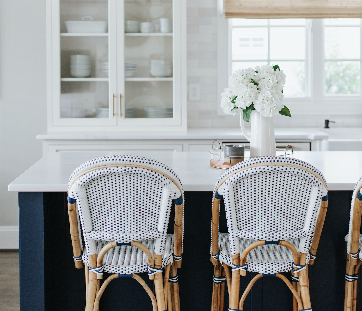 A modern kitchen with a white countertop and navy blue island with woven rattan chairs. The back wall shows white cabinets with glass doors displaying white dishes.